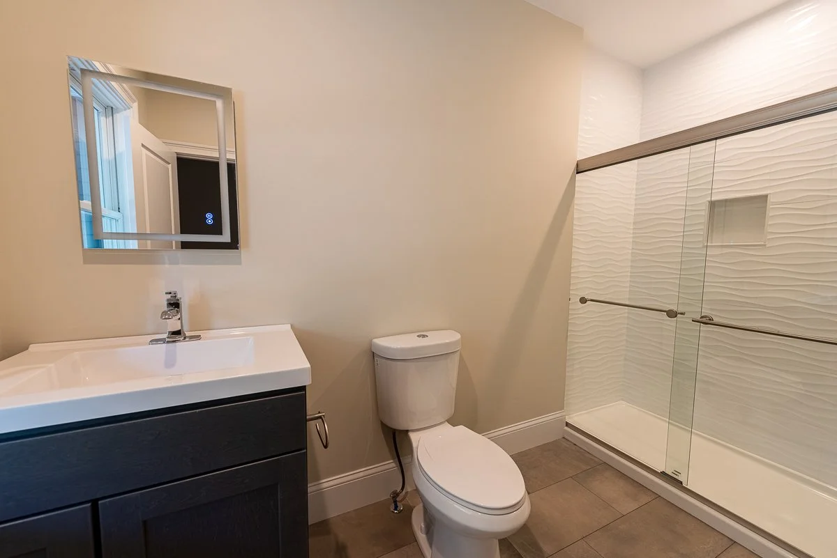 Bathroom with a vanity and mirror, toilet, and a walk-in shower with glass doors and textured white tiles.
