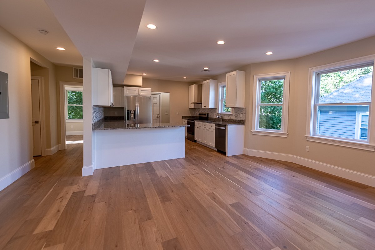 Empty kitchen with hardwood floors, white cabinets, stainless steel refrigerator, and large windows overlooking green trees.