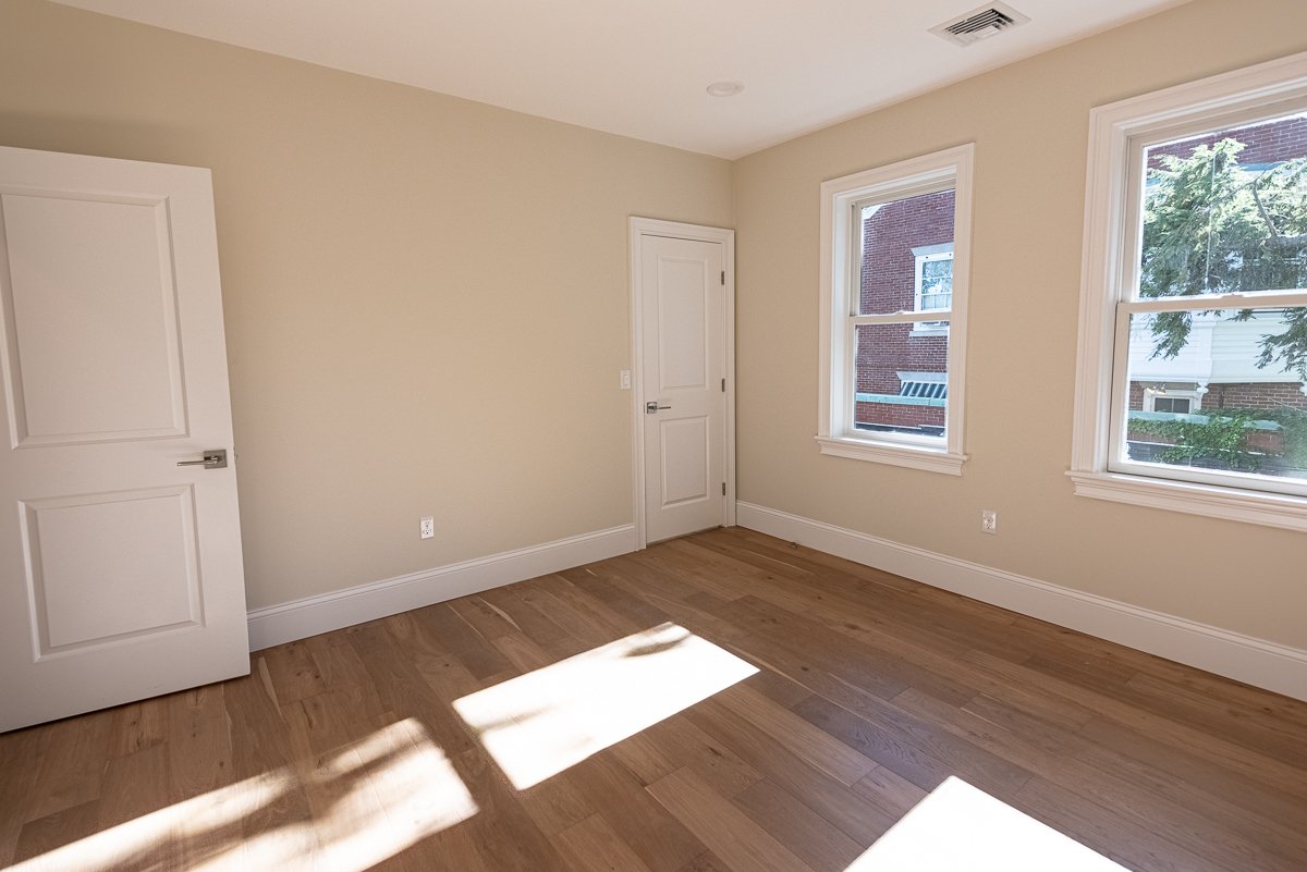 Empty room with beige walls, hardwood floors, two windows letting in natural light, and a white door.