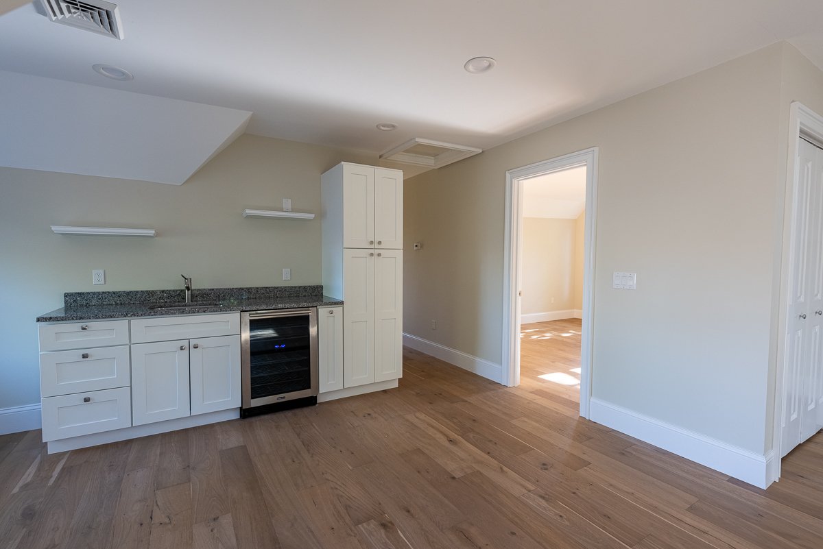 Empty room with a kitchenette, white cabinets, granite countertop, small wine fridge, and an adjoining room with sunlight.