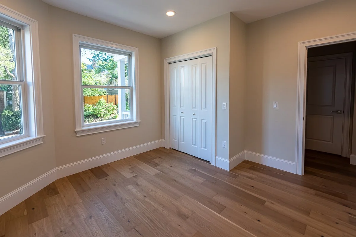 Empty room with three windows showing a view of trees and a fence outside, beige walls, hardwood floor, white baseboards, closet with sliding doors, and doorway leading to another room.