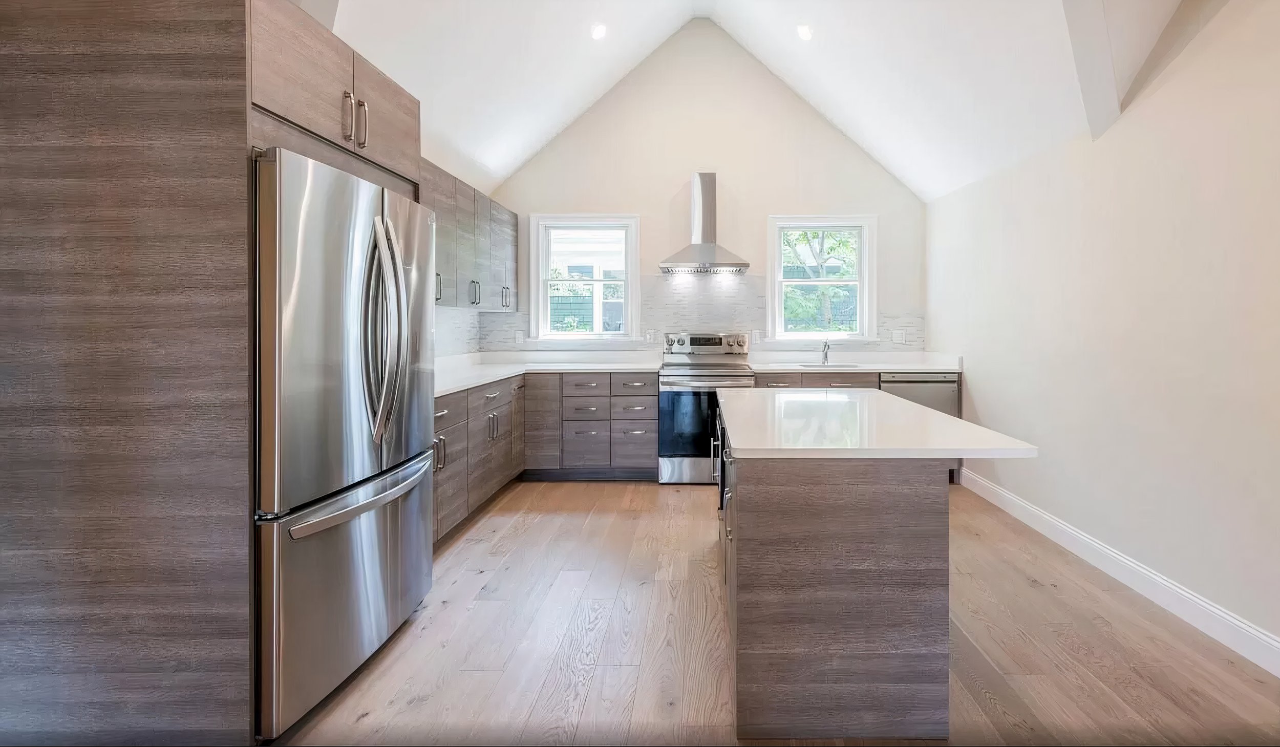 Modern kitchen with wooden cabinets, stainless steel refrigerator, white countertops, two windows, and a stainless steel range hood with stove.
