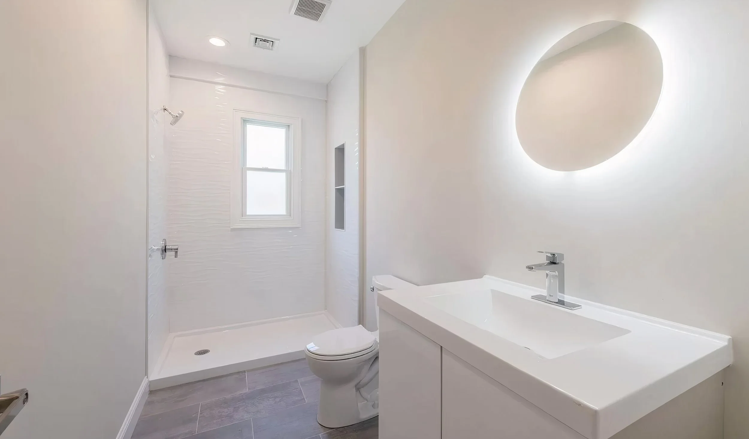 Modern bathroom with white walls, a window, a shower area with a rain shower head, a toilet, and a white vanity with a rectangular sink and a silver faucet. There is a circular mirror with backlit lighting on the wall.