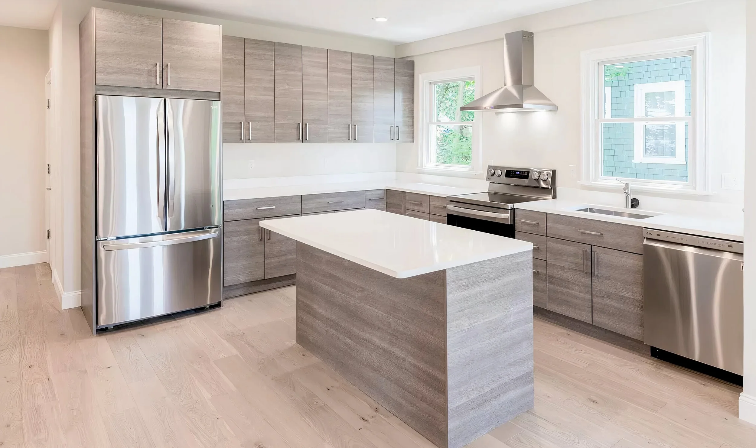 Modern kitchen with gray cabinets, stainless steel refrigerator, stove, and dishwasher, white countertops, and two windows providing natural light.