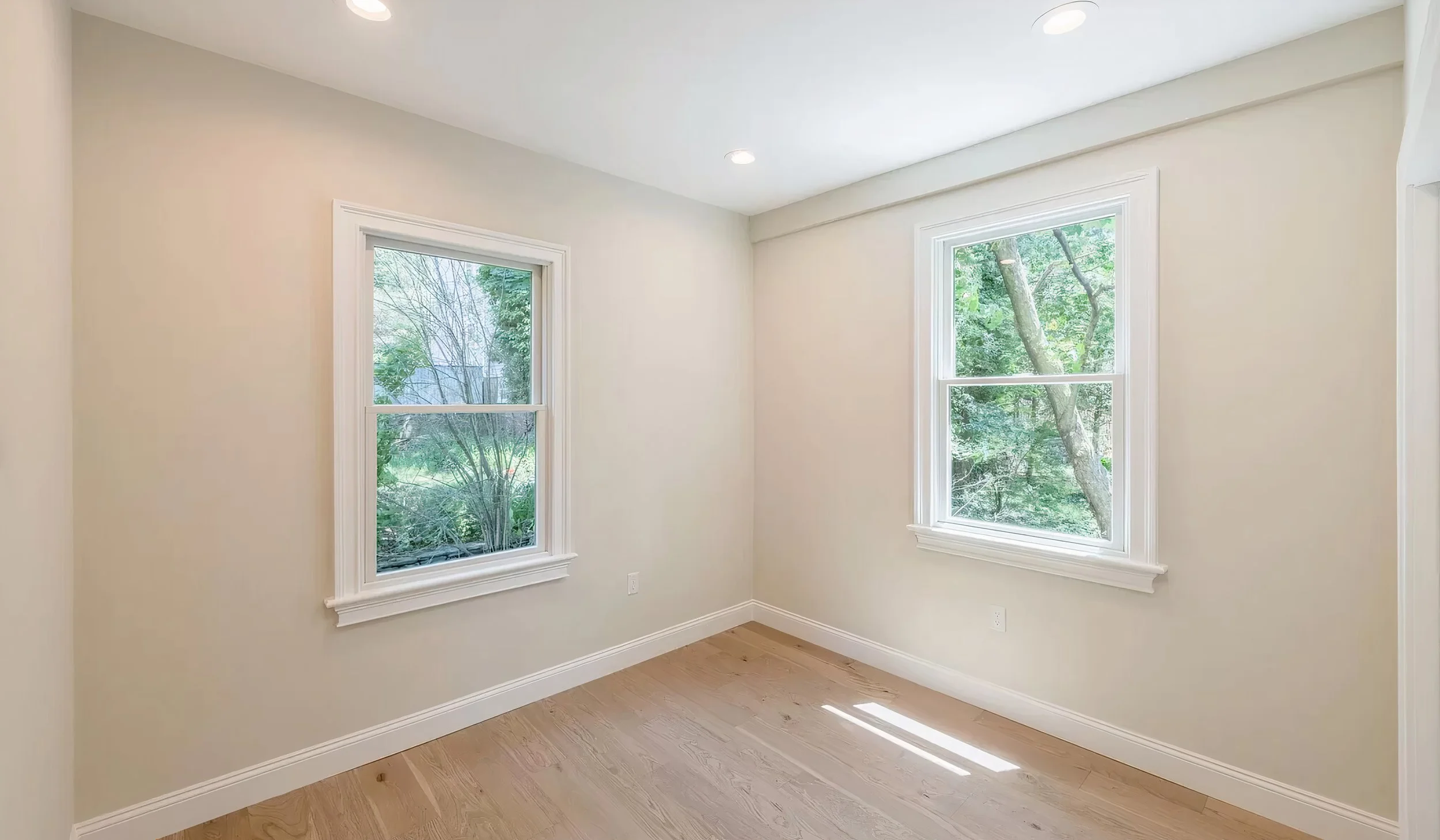 Empty beige room with two large windows showing green trees outside, hardwood floors, and white trim.