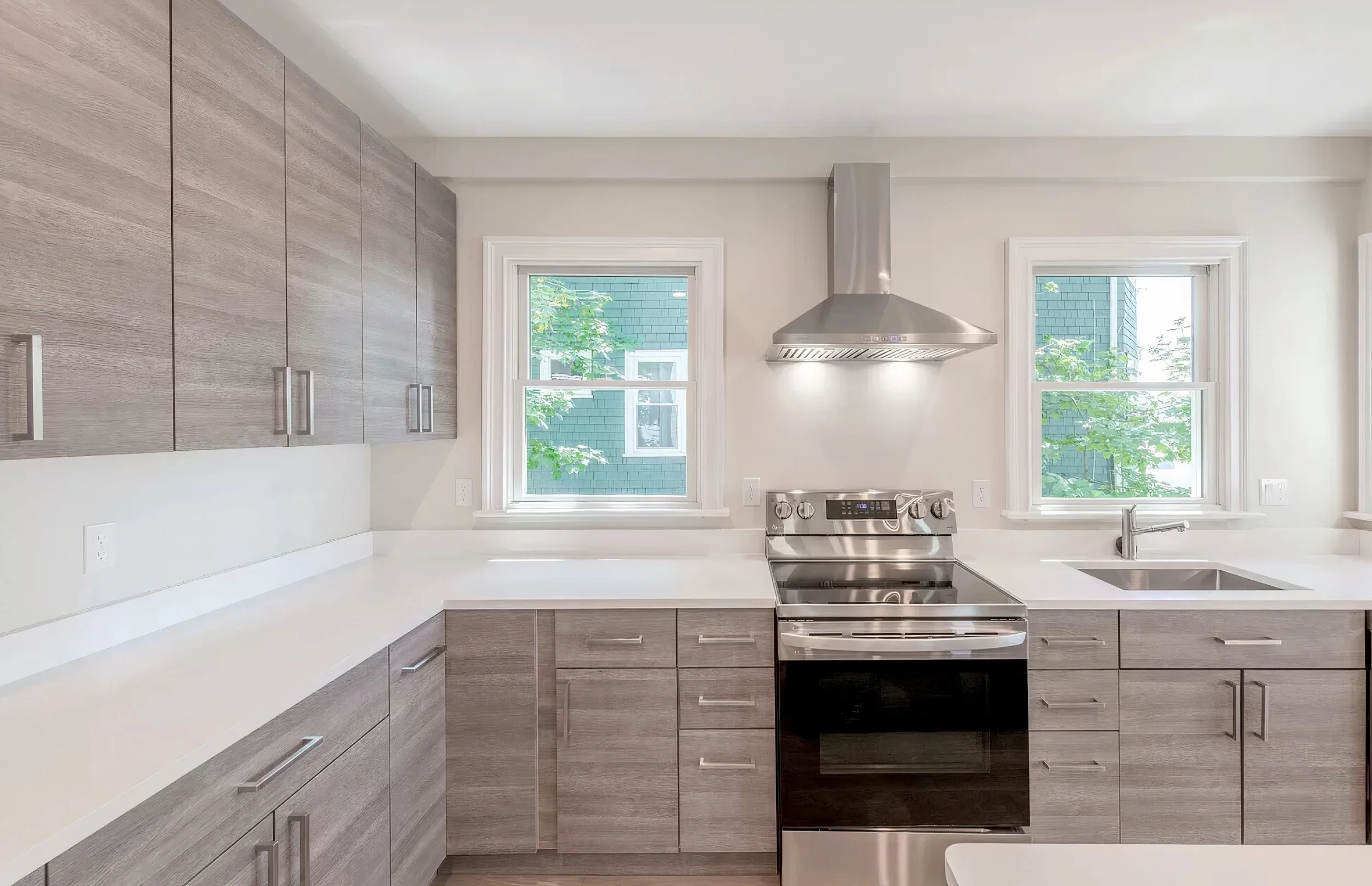 Modern kitchen with gray wood cabinets, white countertops, a stainless steel stove, stainless steel range hood, and two windows showing green trees outside.