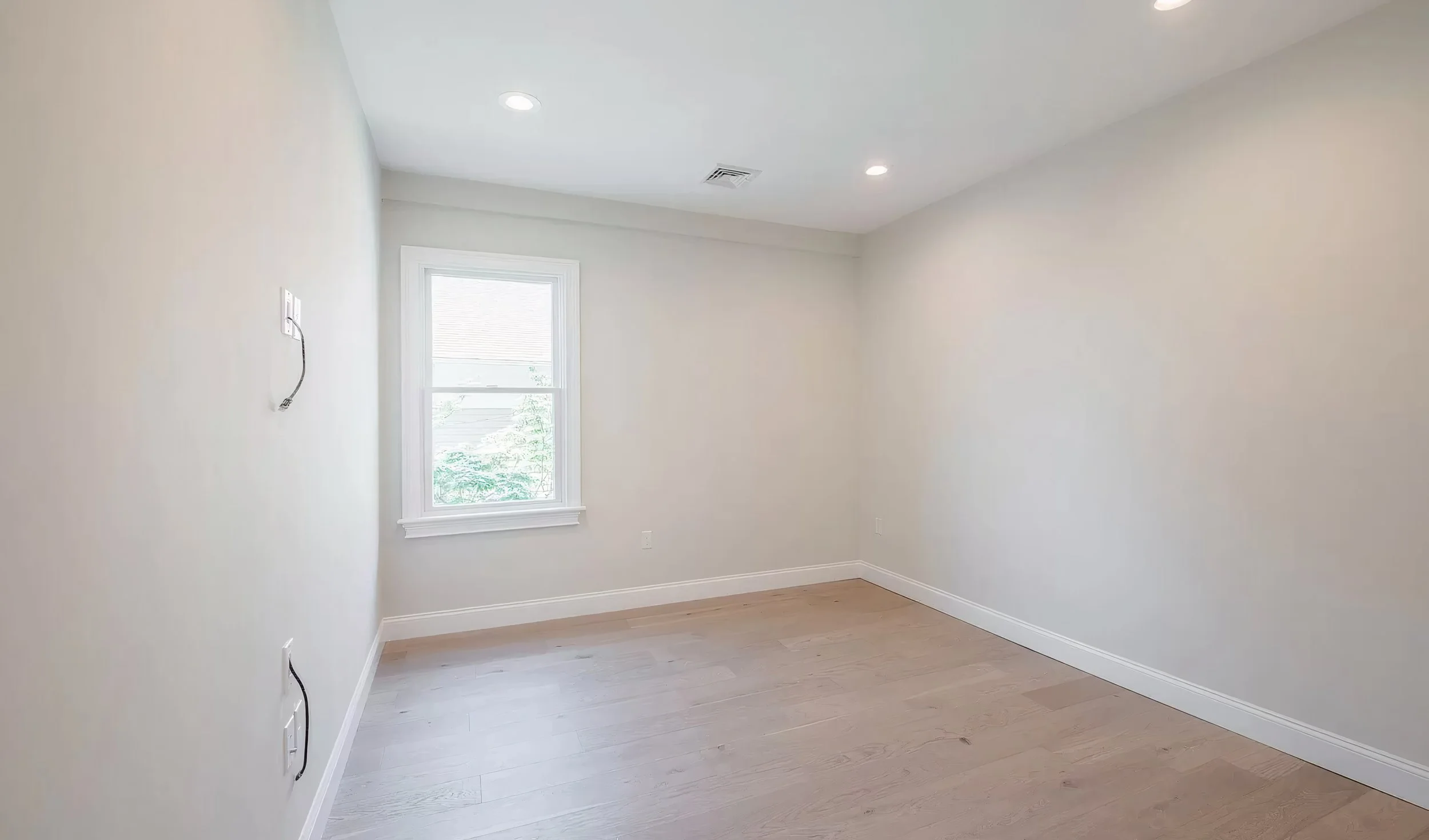 An empty room with white walls, a window with blinds, recessed lighting, and light wood flooring.