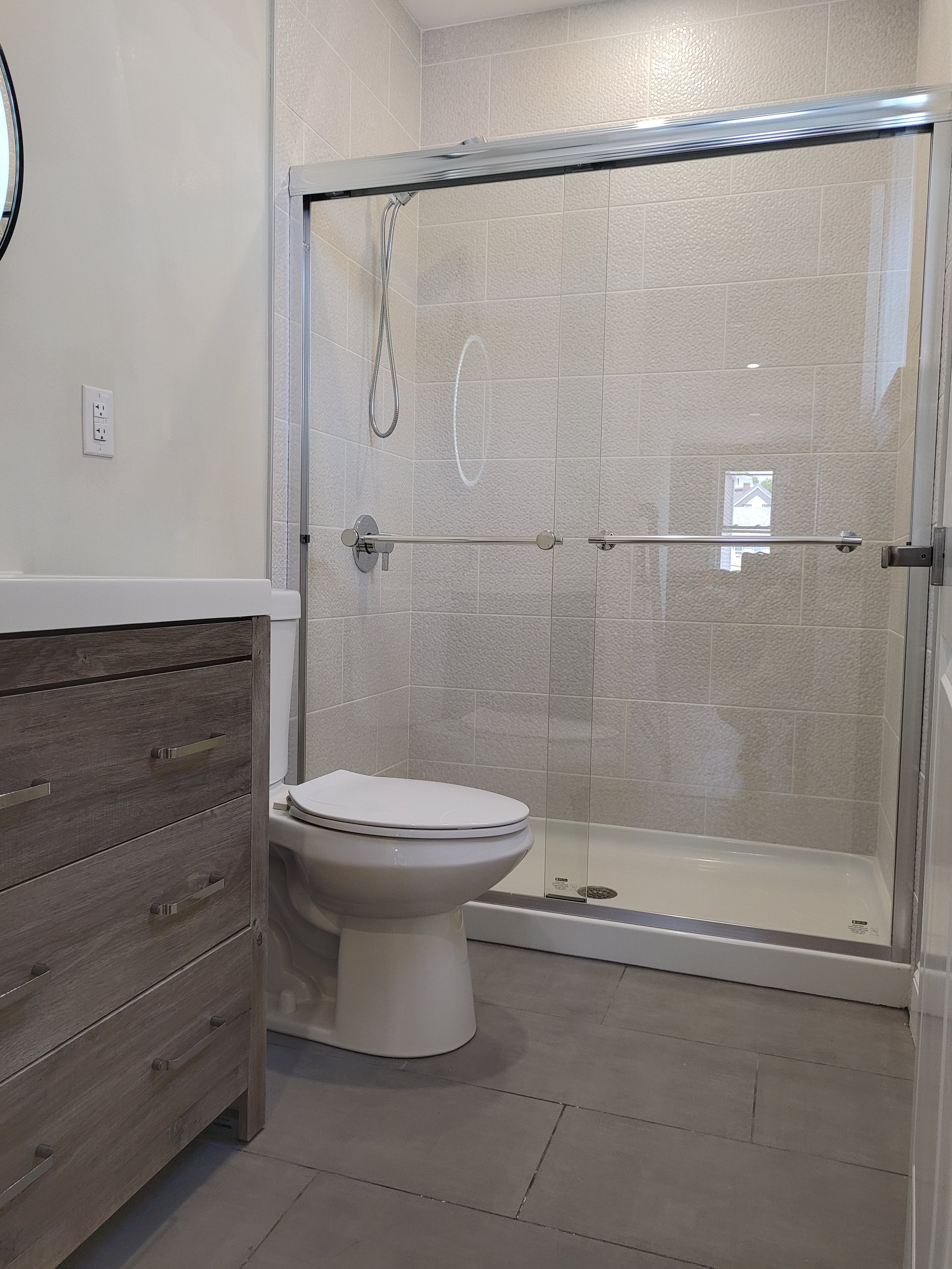 Bathroom with a glass shower enclosure, a toilet, a wooden vanity with drawers, and tiled walls and floor.