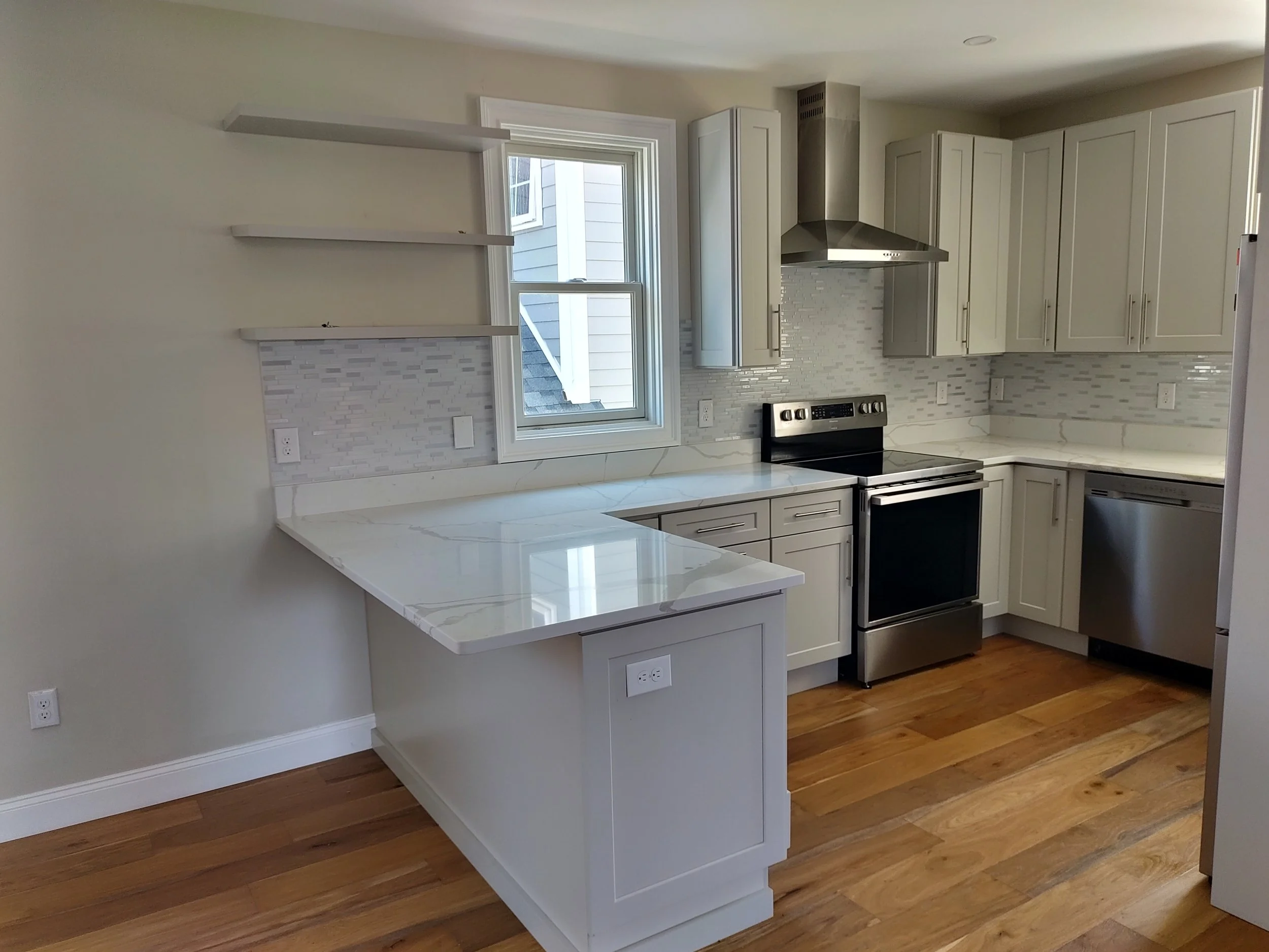 Modern kitchen with white cabinetry, stainless steel appliances, a marble countertop, a window overlooking a house exterior, and hardwood flooring.