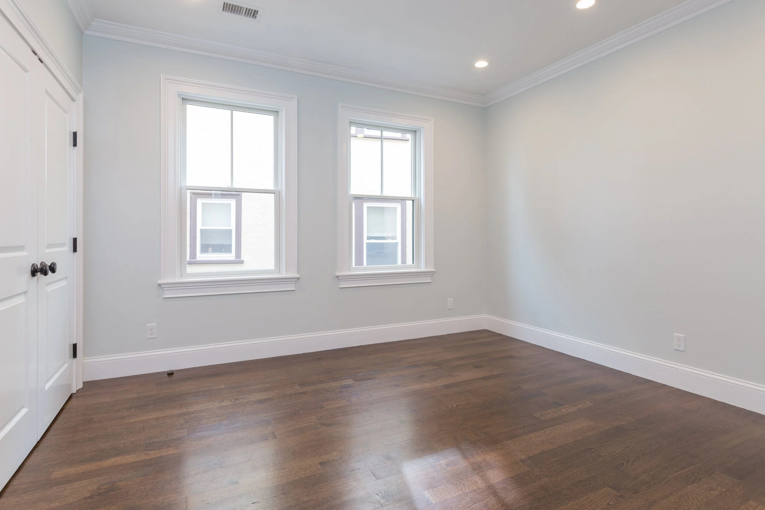 Empty room with white walls, two windows, dark hardwood flooring, and a white door.