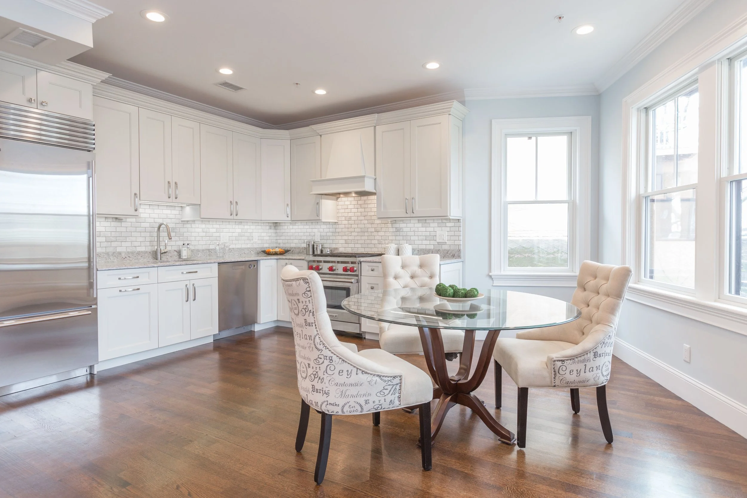 A modern white kitchen with hardwood floors, white cabinets, a brick backsplash, stainless steel appliances, and a dining area with a round glass table and upholstered chairs.