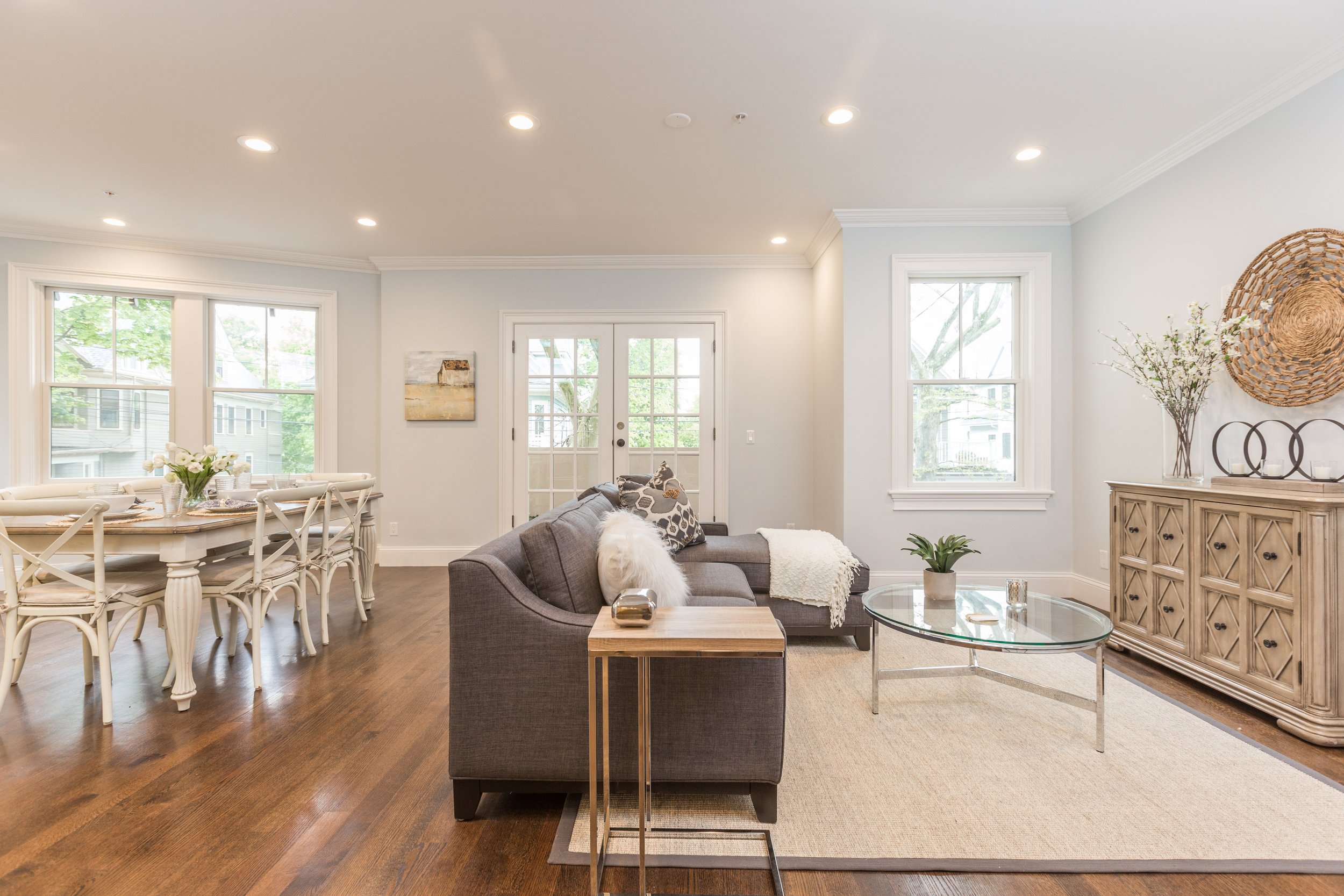 Bright living room with white walls, hardwood floors, large windows, and glass double doors, decorated with a gray sofa, coffee table, and decorative items like plants, artwork, and woven wall hanging.