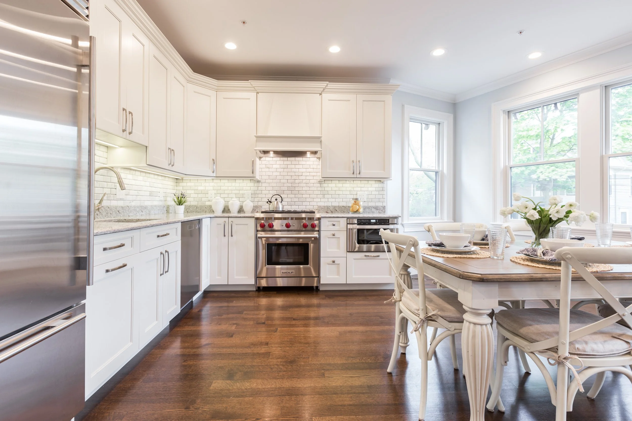 Bright modern kitchen with white cabinets, stainless steel appliances, dark hardwood floors, and a dining area with a table set for four, decorated with flowers and placemats, surrounded by large windows.