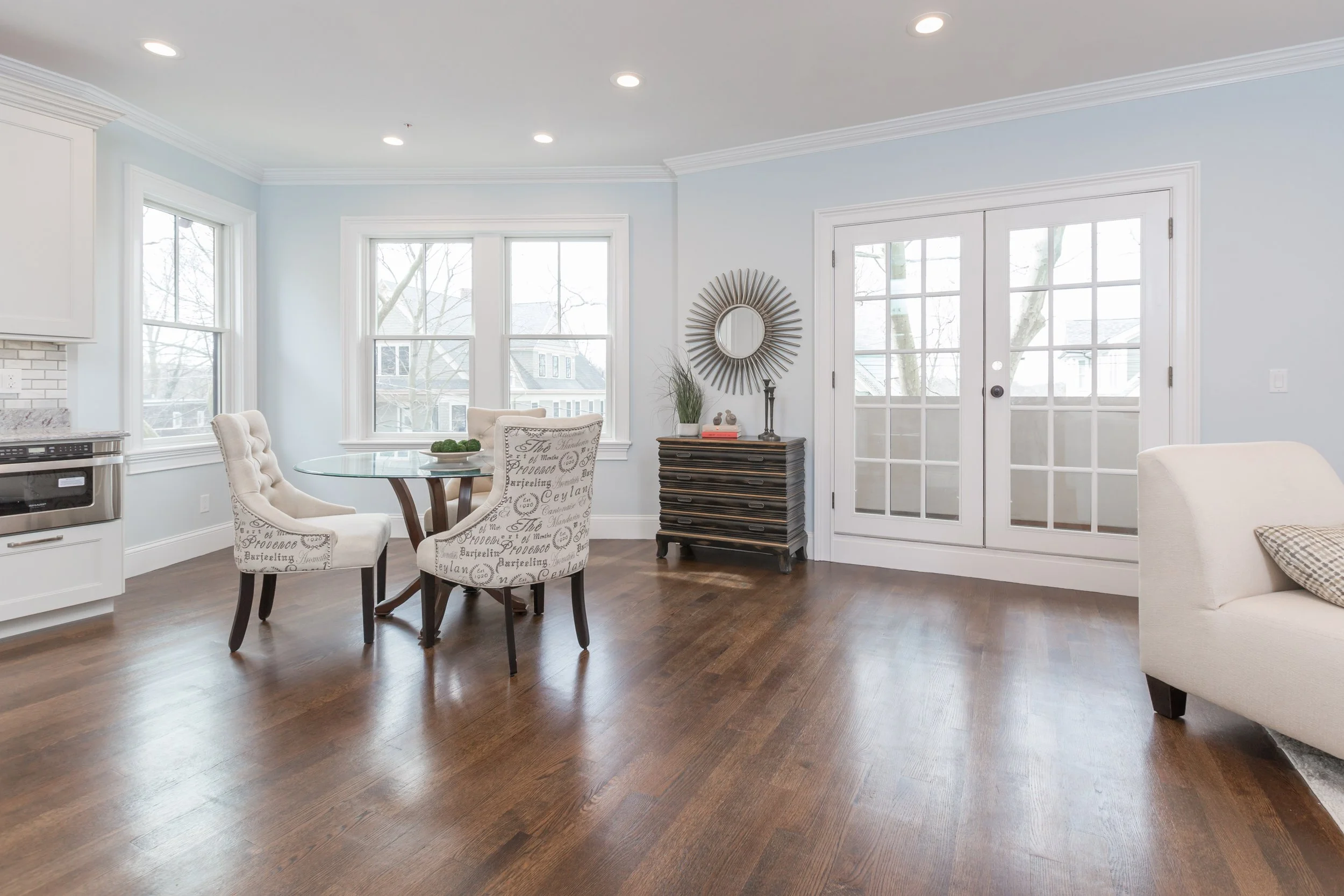 A bright living room with hardwood flooring, large windows, white walls, and a glass dining table with upholstered chairs. There is a small black chest of drawers with decorative items, a round mirror, and a sliding glass door leading outside.