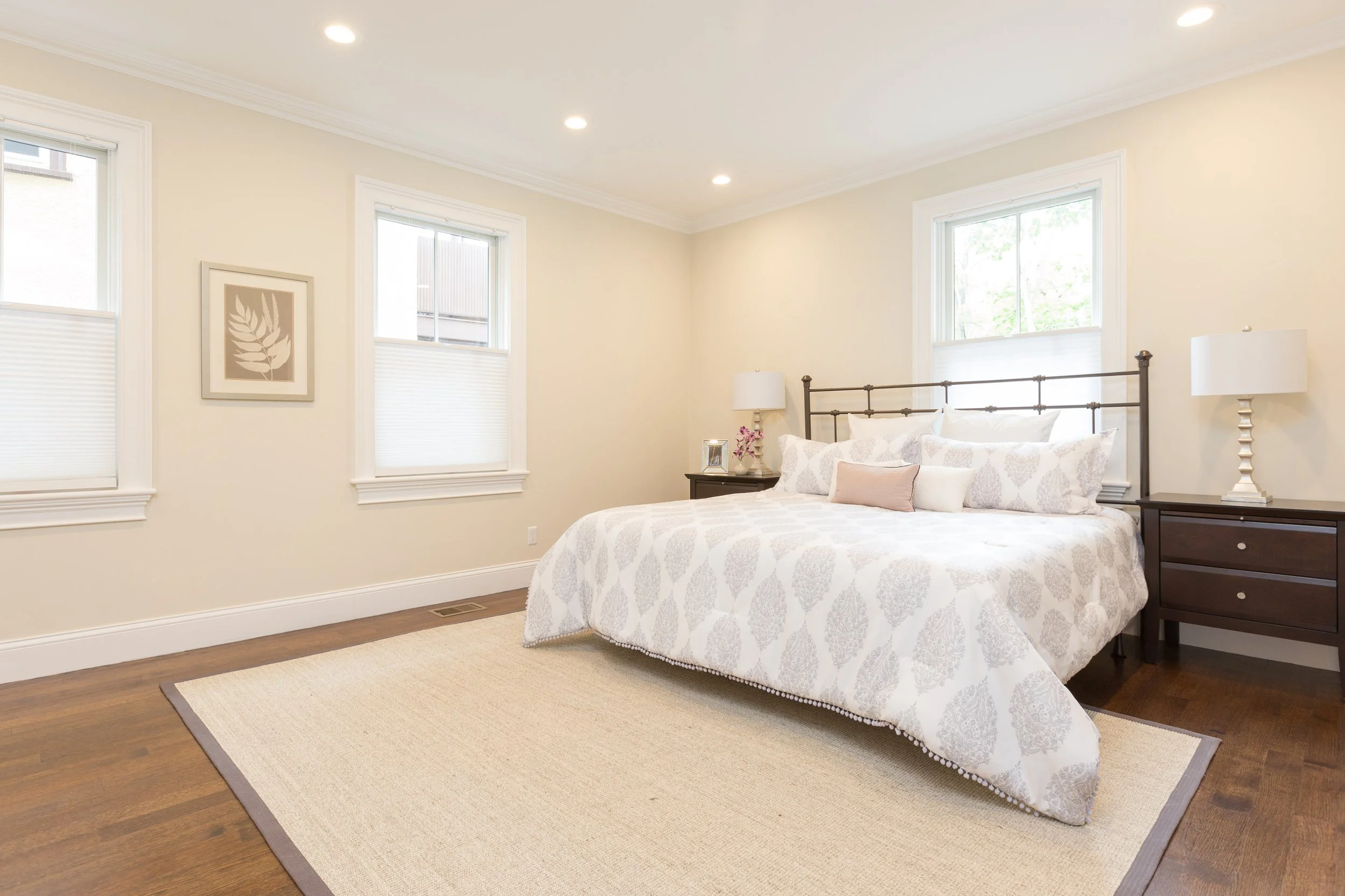 Bedroom with three windows, a bed with white patterned linens, two bedside tables with lamps, a picture on the wall, and a beige area rug on wooden floors.