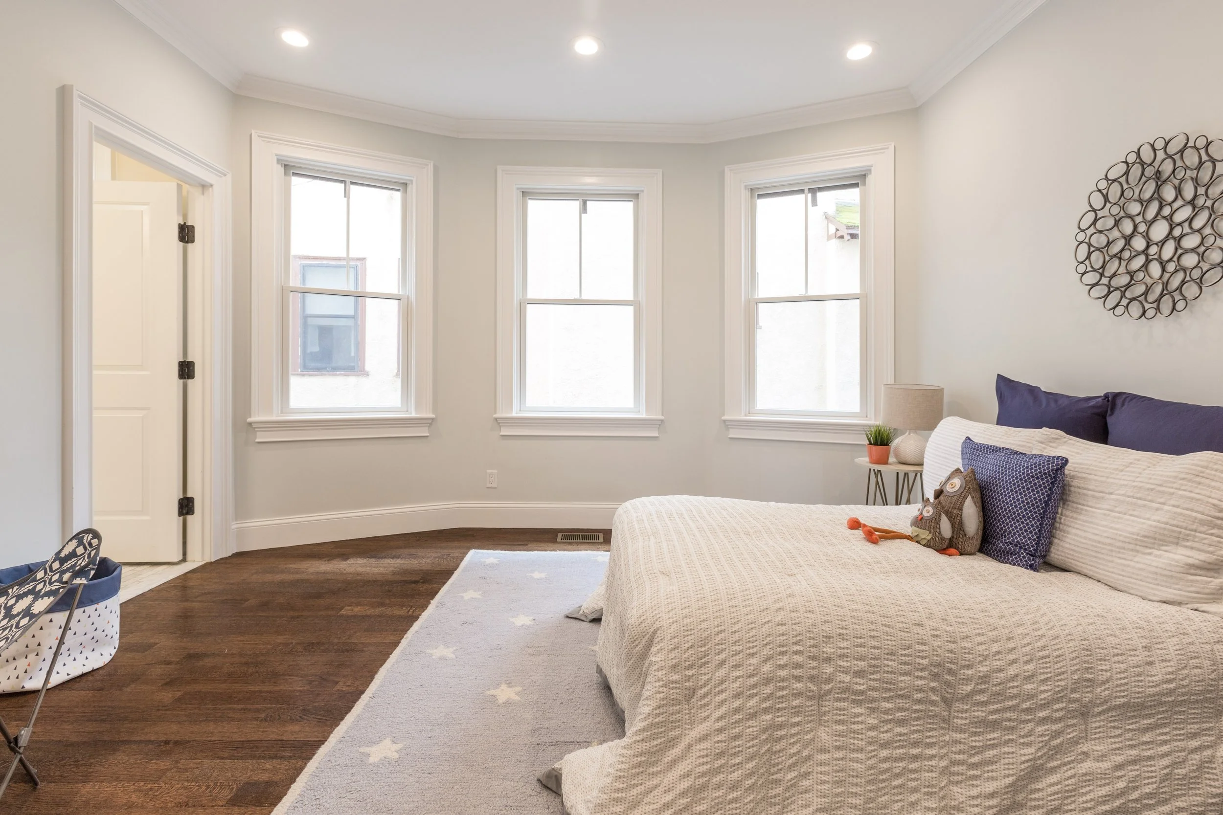 A cozy bedroom with three large windows, crown molding, wooden floors, a bed with a textured cream-colored blanket, and decorative pillows. There is a nightstand with a lamp, a small plant, and stuffed animals.