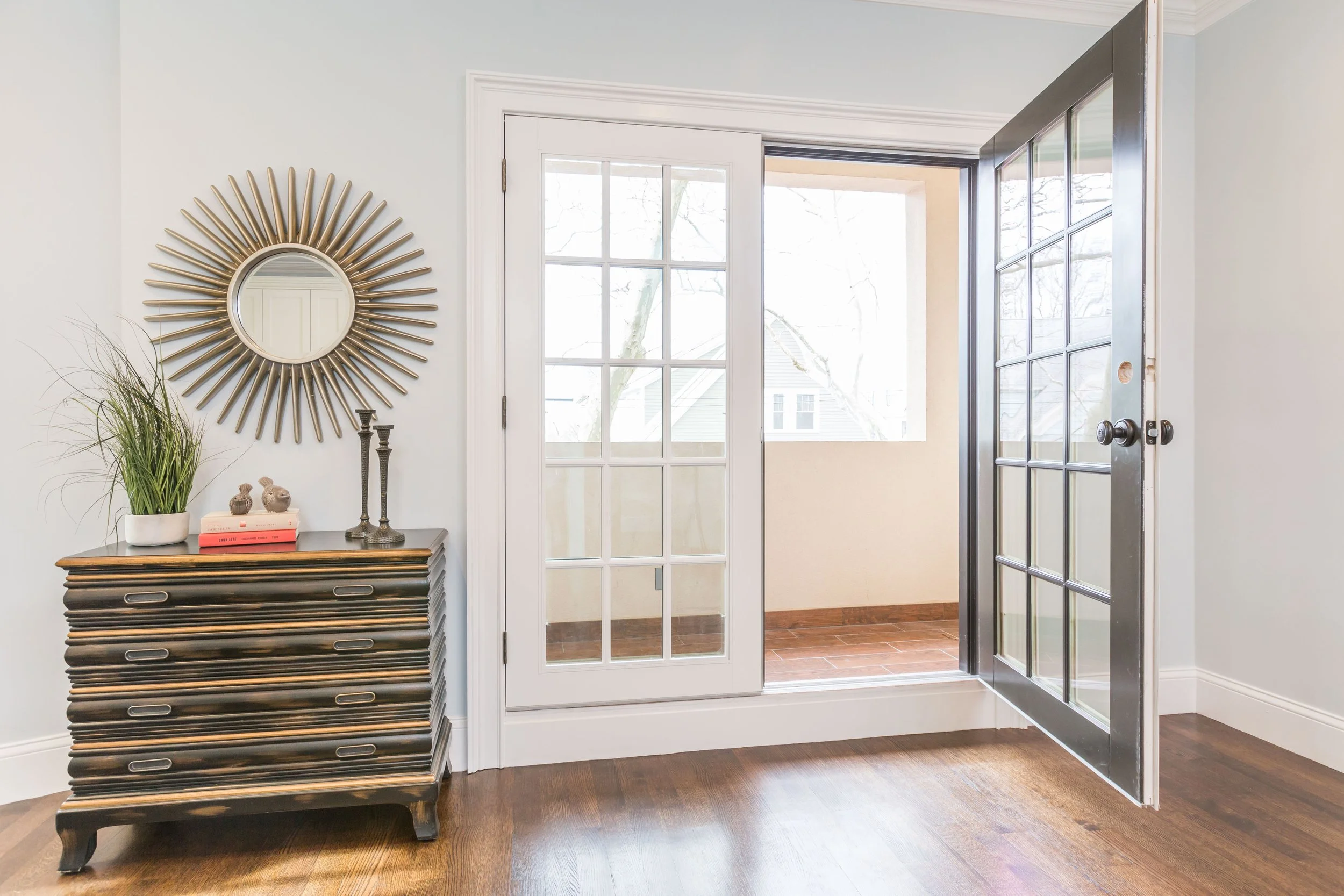 Entrance area with a front door made of glass and a metal frame, leading to an outdoor porch with a tree visible outside. A decorative black dresser with multiple drawers is decorated with a potted plant, two small ceramic animal figurines, and a few