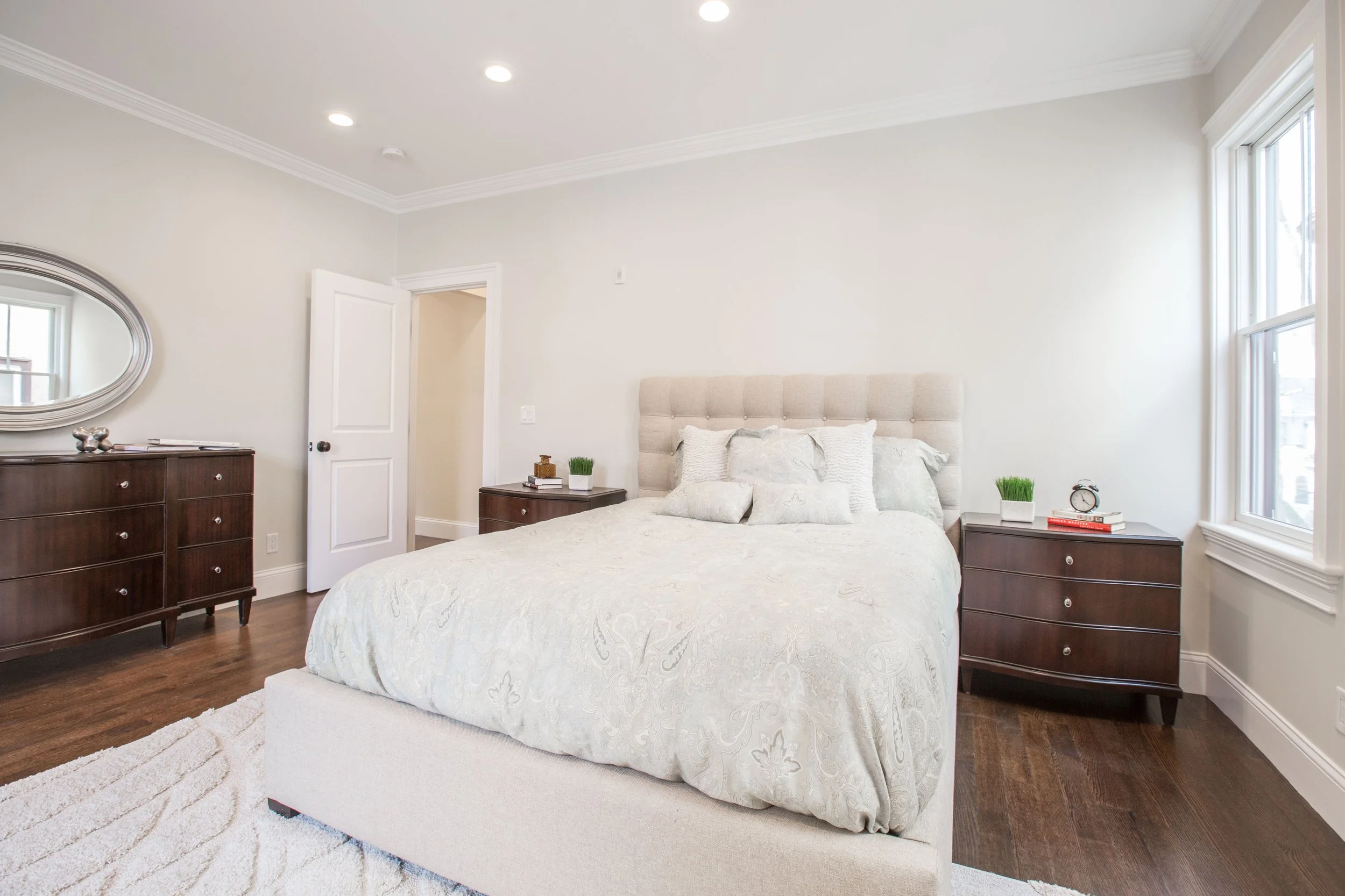 A bedroom with a large bed, matching nightstands, and a dresser with a mirror. The room has white walls, hardwood floors, and a window with natural light.