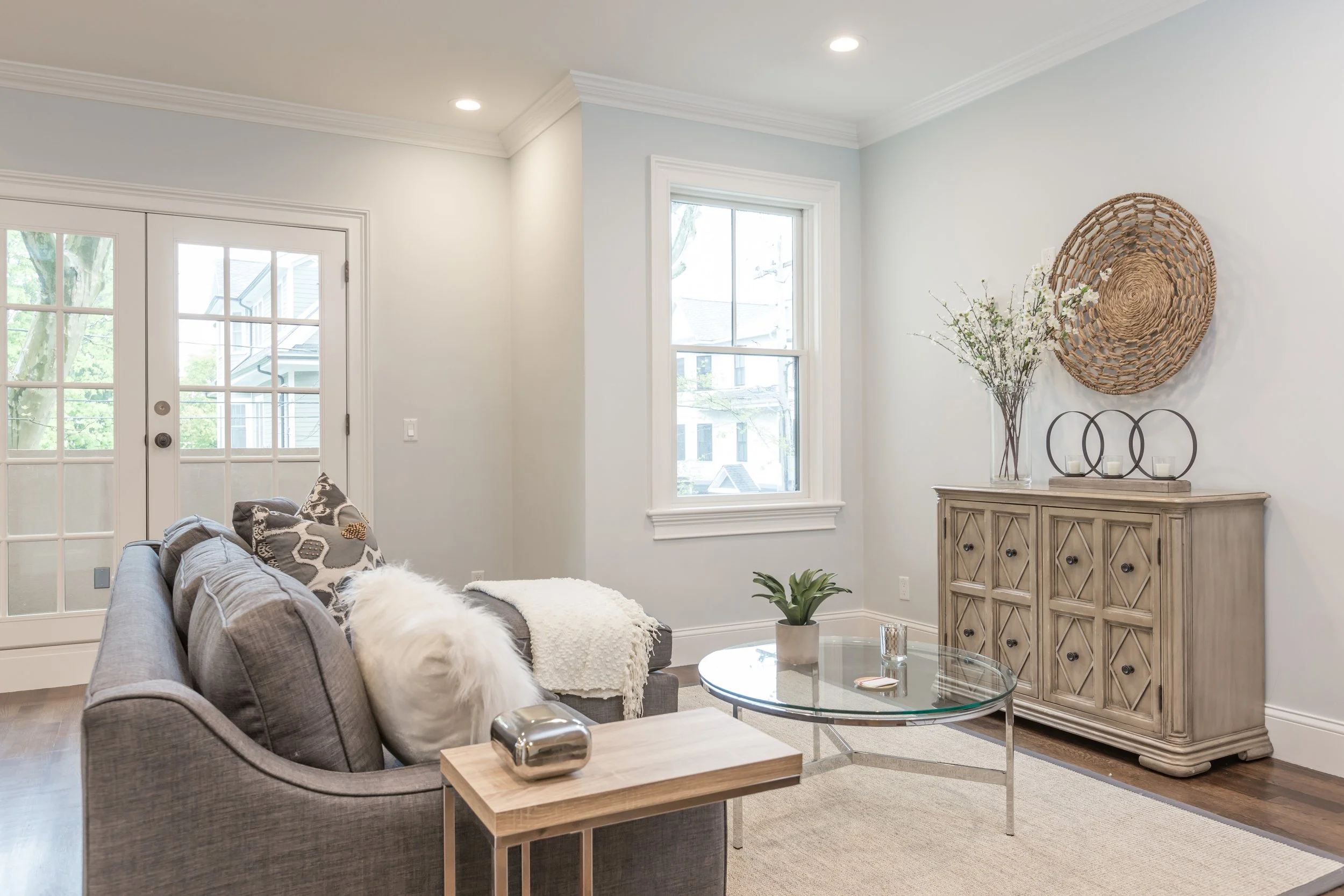 Living room with gray sofa, throw pillows, glass coffee table, side table, wooden cabinet, wall decor, and large windows letting in natural light.