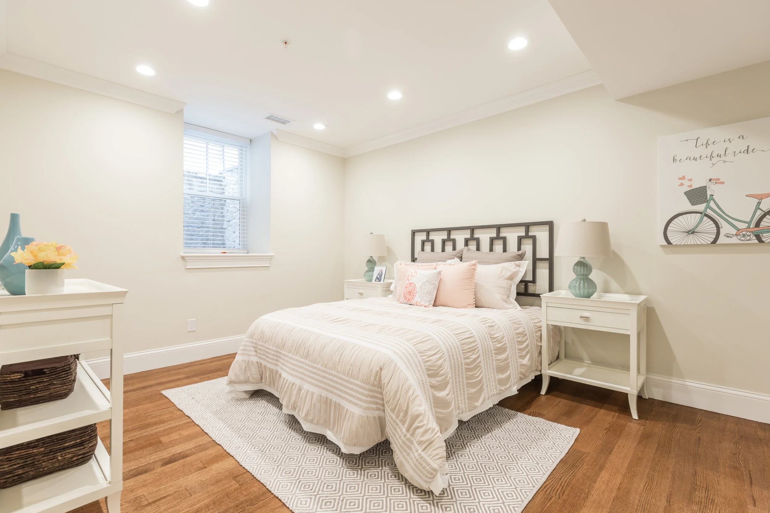 A bedroom with a white bed, nightstands, lamps, and artwork, featuring a window with blinds and hardwood flooring.