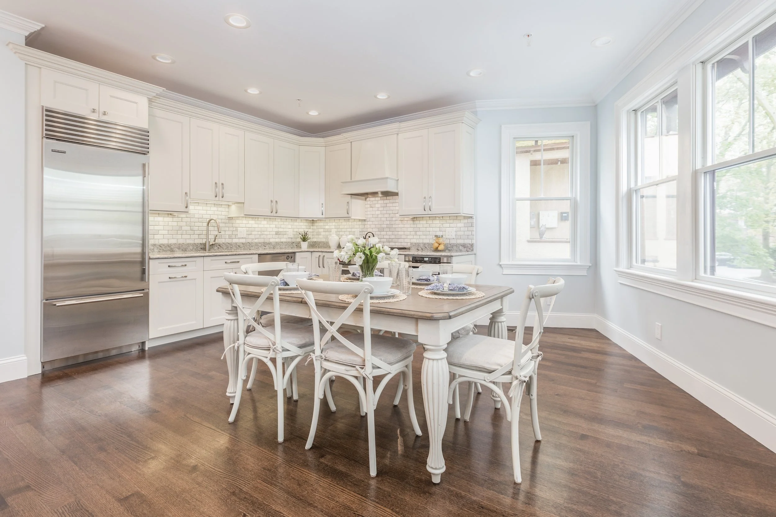 Bright kitchen with white cabinets, granite countertops, stainless steel appliances, and a dining table set for six with white chairs and a vase of white flowers.