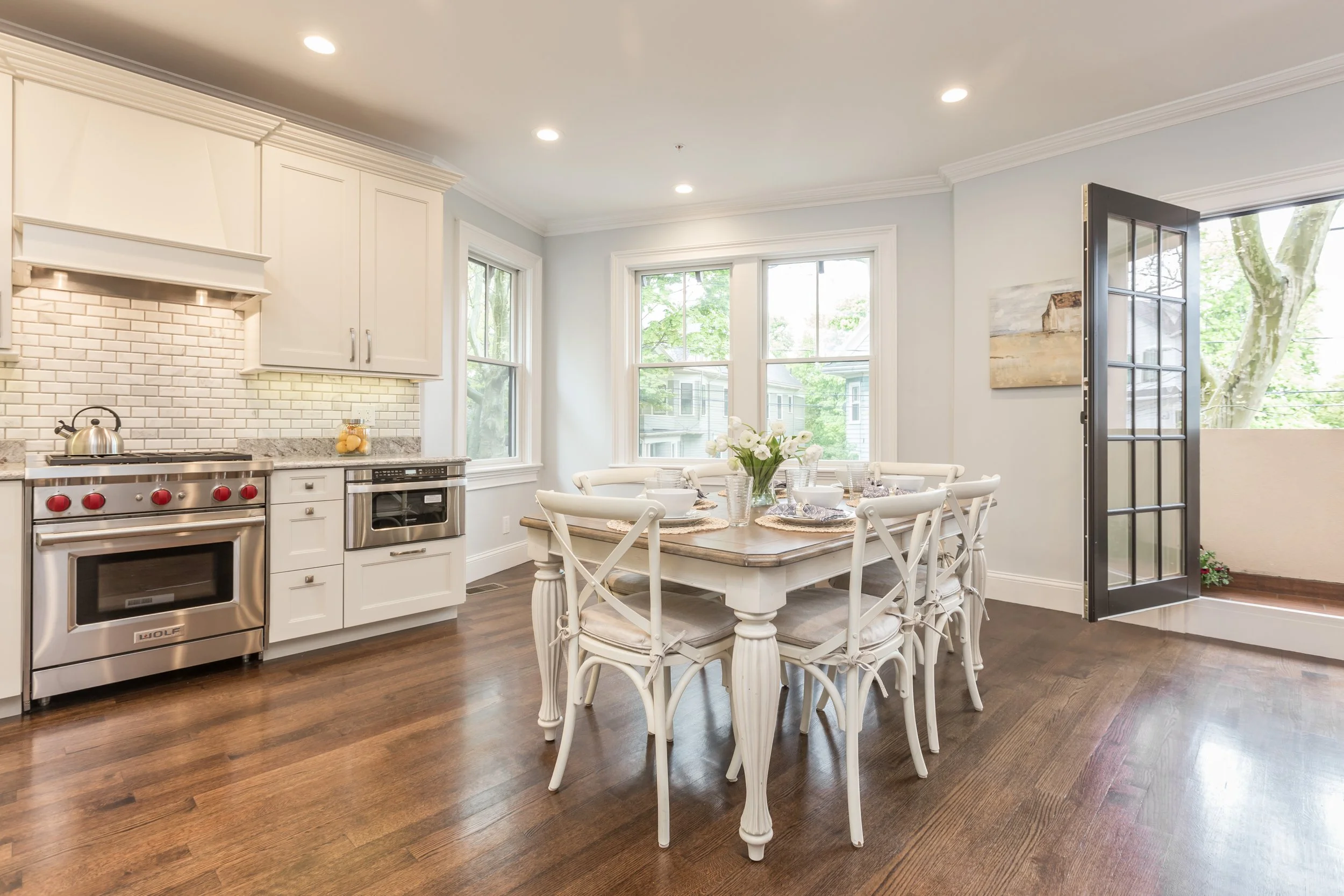 Bright kitchen and dining area with white cabinetry, stainless steel appliances, a round dining table with white chairs, large windows, and an open door leading outside, with hardwood floors.