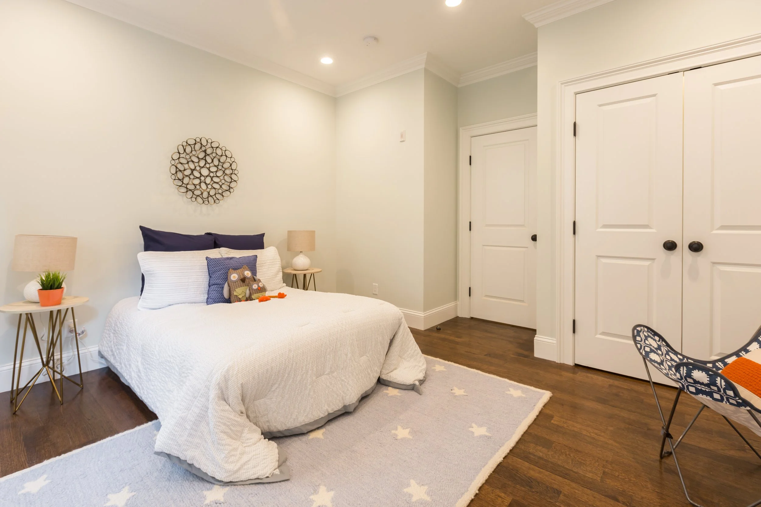 A bedroom with a bed, two side tables with lamps, a decorative wall hanging, a plant, and a lounge chair, with white walls and wood floors.