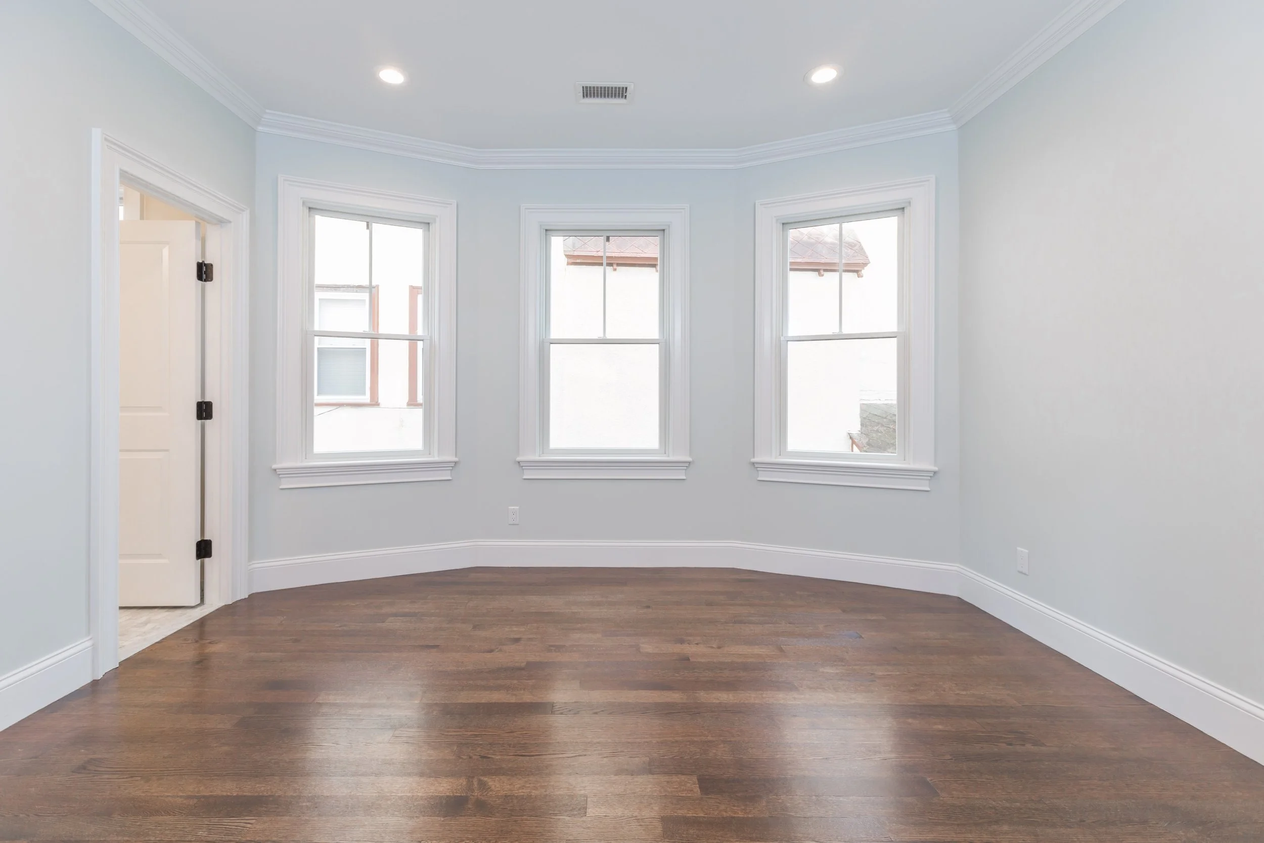 Empty room with white walls, crown molding, three large windows, and dark hardwood floors.