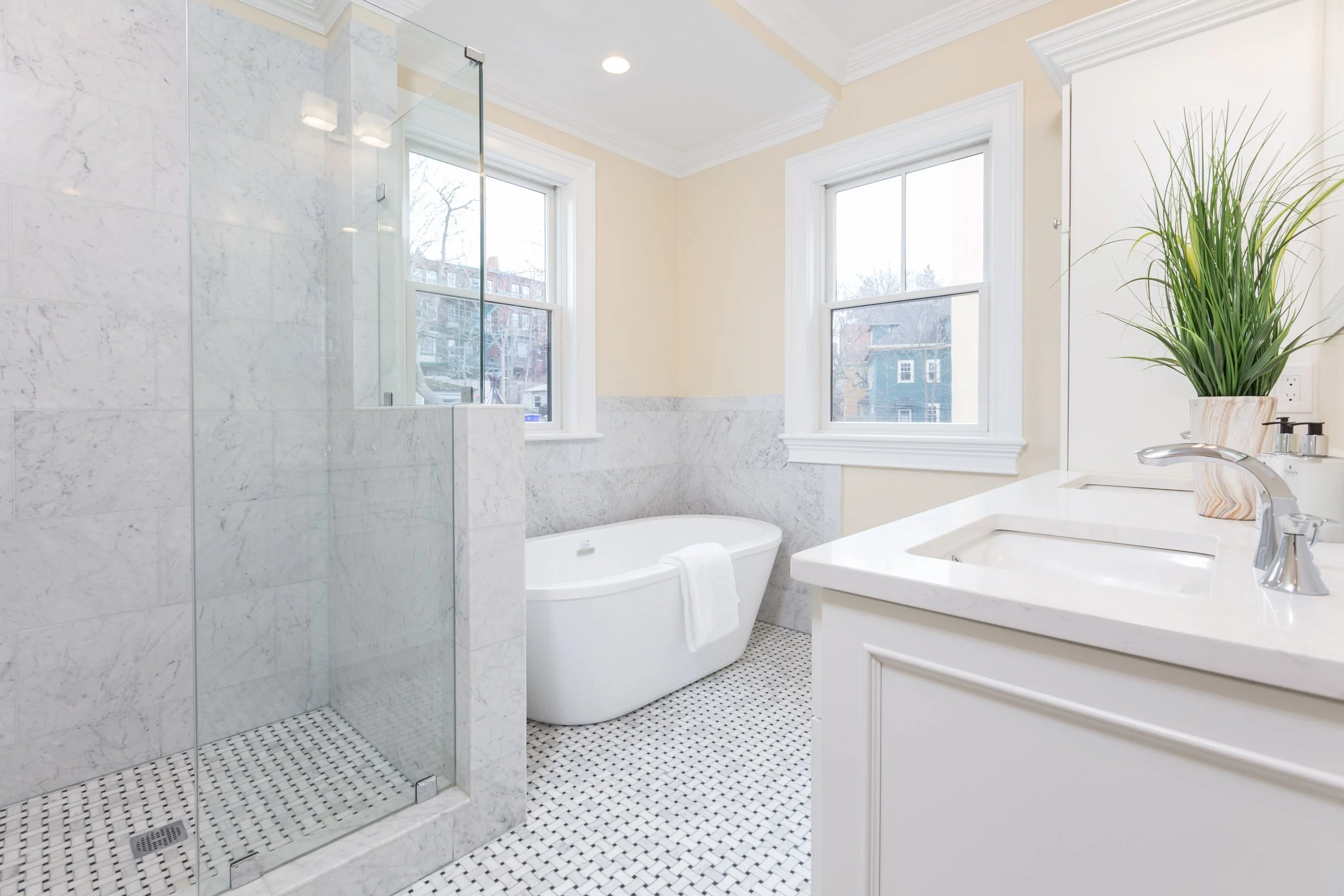 Bright bathroom with two large windows, marble-tiled walls, a white bathtub, a glass shower enclosure, a white vanity with a sink, a potted plant, and a patterned tile floor.