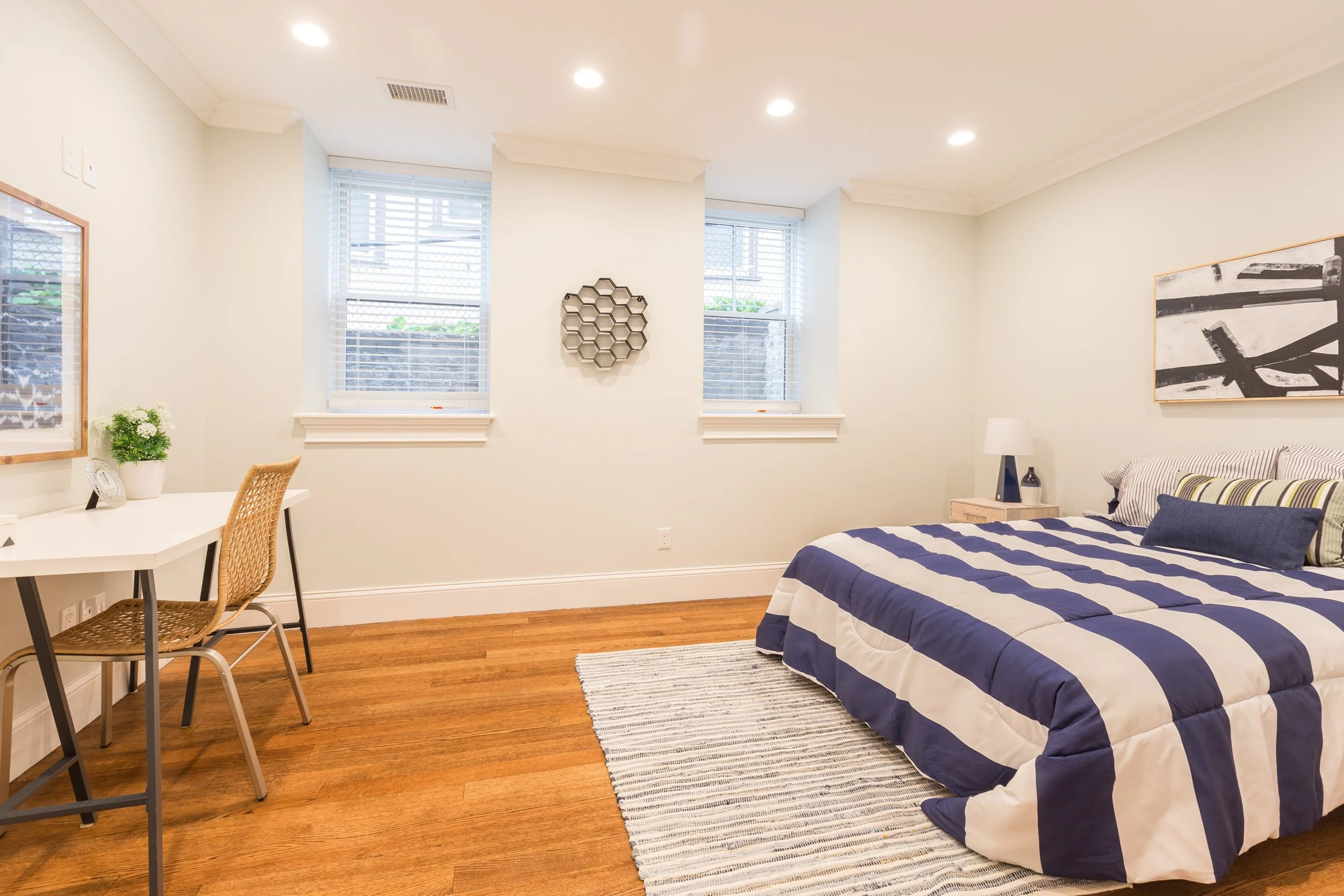 A bedroom with white walls, hardwood floors, and a bed with a striped navy blue and white comforter. There are two large windows with white blinds and decorative frames. The room has modern ceiling lights, wall art, a small side table with a lamp, an