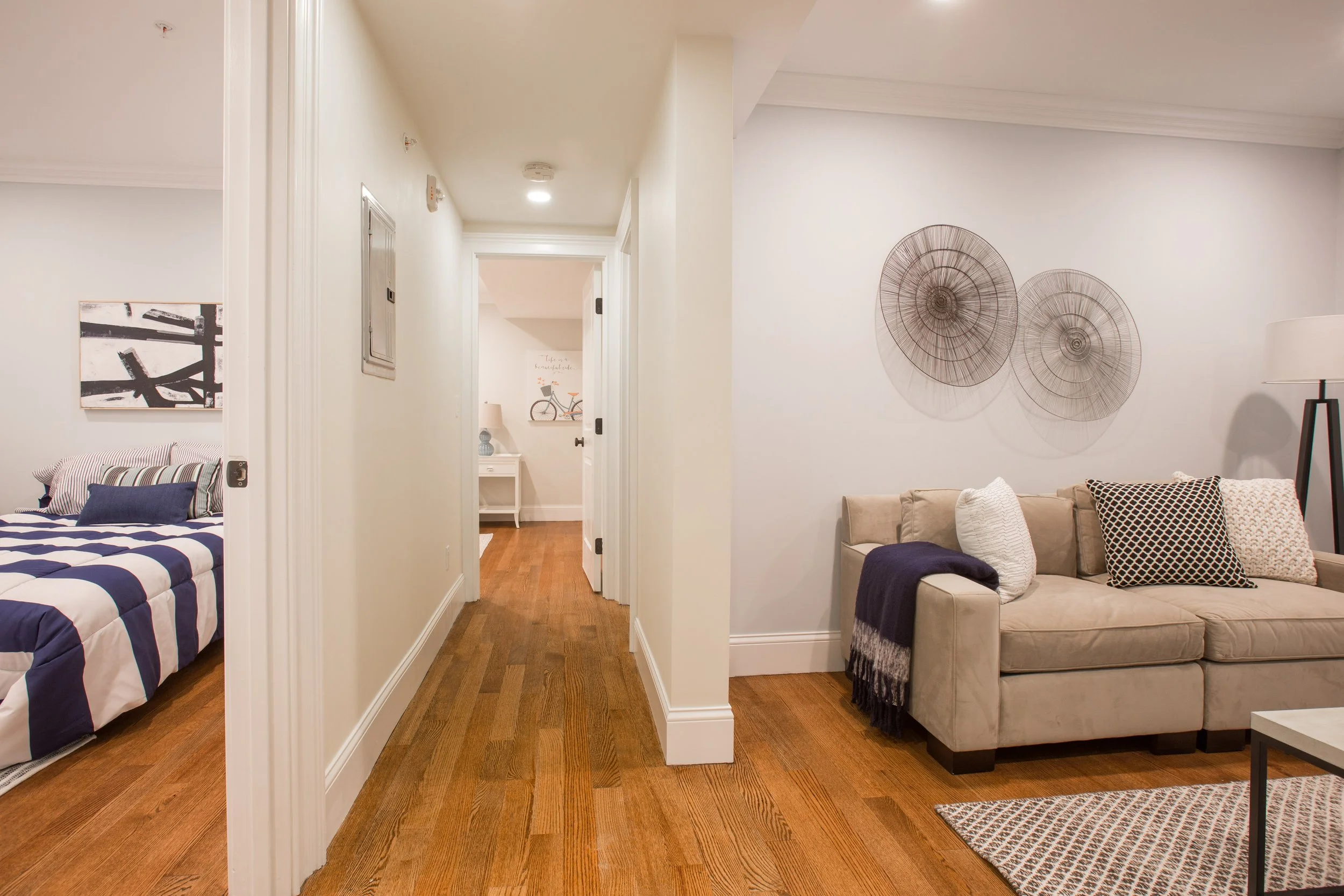 Interior view of a hallway with hardwood flooring, leading to a bedroom on the left with a bed and artwork on the wall, and a living room area on the right with a beige sofa, decorative pillows, and a wall sculpture.