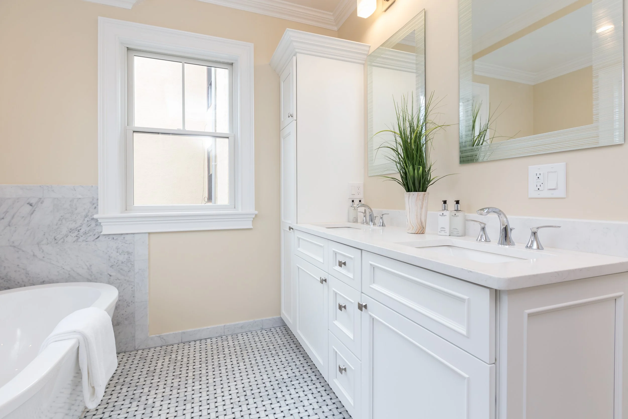 A bathroom with a white vanity, two sinks, and a large mirror. There is a potted green plant on the counter, and a window letting in natural light. The wall is painted a light beige, and the floor has a black and white tile pattern.