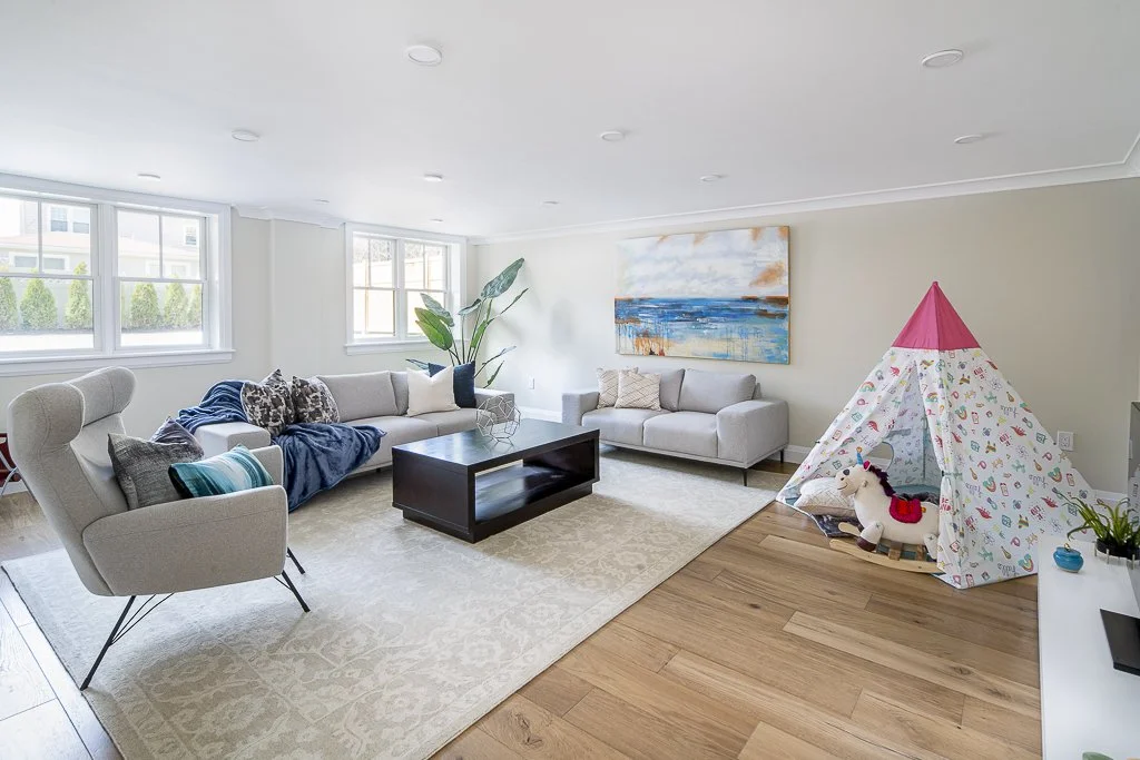 Living room with light-colored sofas, a beige armchair, a black coffee table, large windows, a plant, wall art depicting a beach scene, and a children's play tent with a plush horse toy.