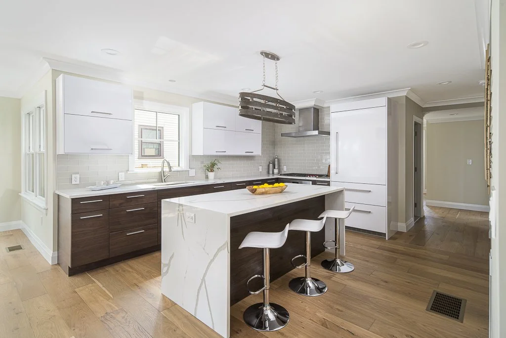 Modern kitchen with white and dark wood cabinets, a white marble island with seating, a window above the sink, chrome appliances, and hardwood floors.