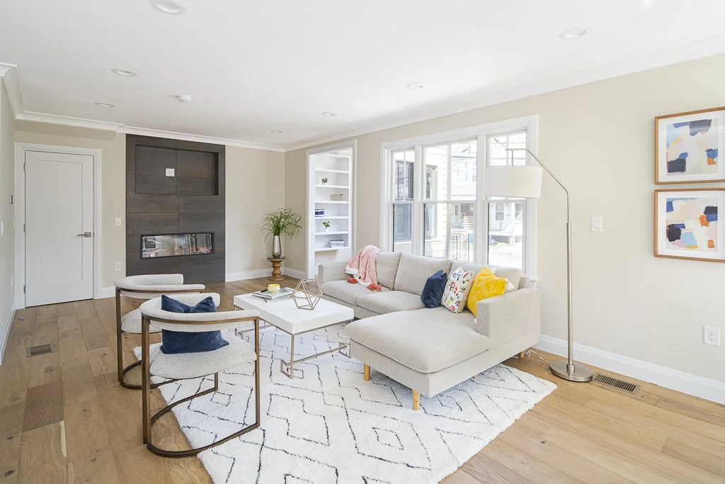 Bright living room with beige sofa, white coffee table, two white chairs with wooden arms, abstract artwork on the wall, fireplace, and large windows letting in natural light.