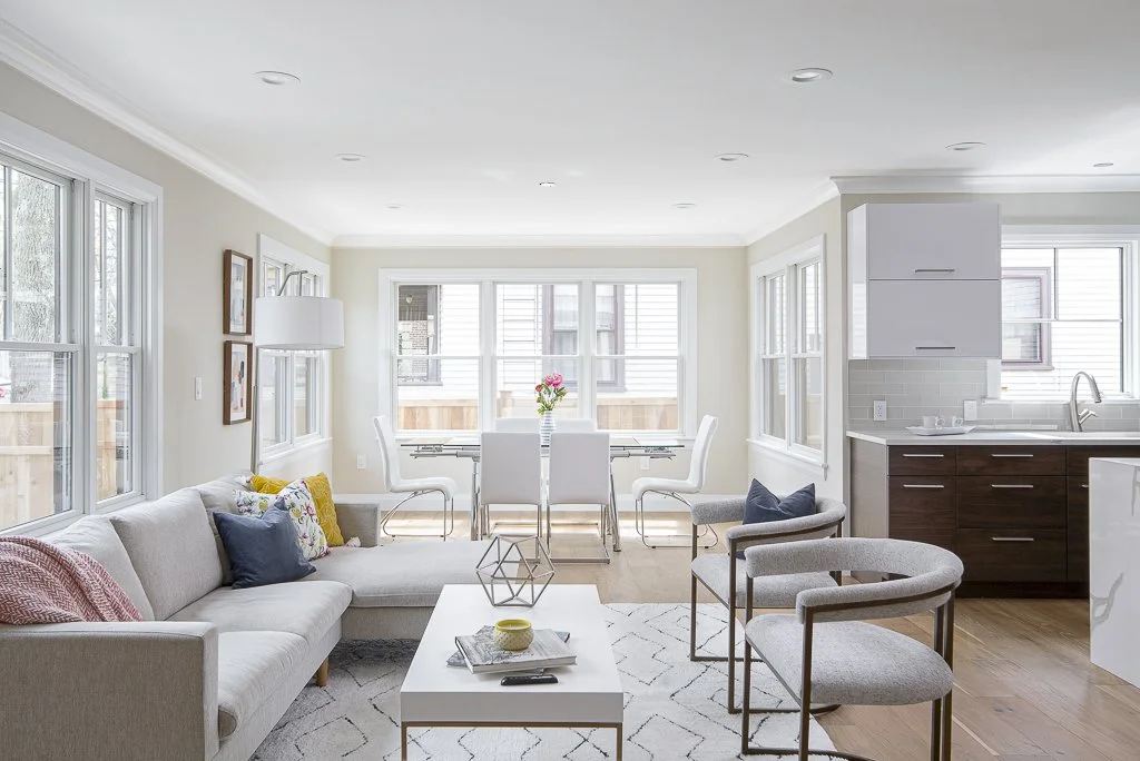 Bright, open living room with large windows, white walls, a sectional sofa with colorful and navy pillows, two modern armchairs, a white coffee table, and a dining area with a table, white chairs, and a vase of pink flowers. The kitchen has dark wood