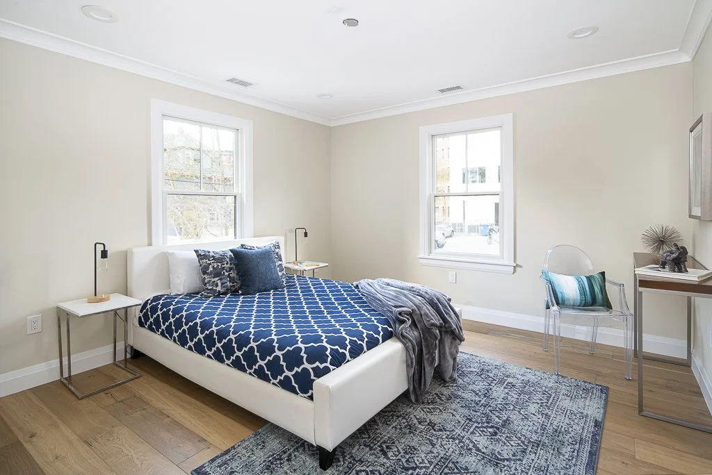 A bright bedroom with a white bed, blue patterned bedding, two windows, a transparent chair with a teal pillow, a small desk, and a gray throw blanket.