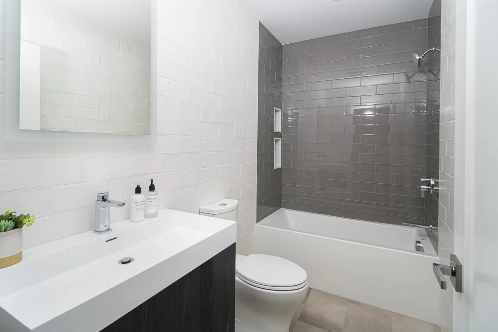 Modern bathroom with white tiled walls, gray tiled shower area, white bathtub, toilet, and a vanity with a white sink and mirror.