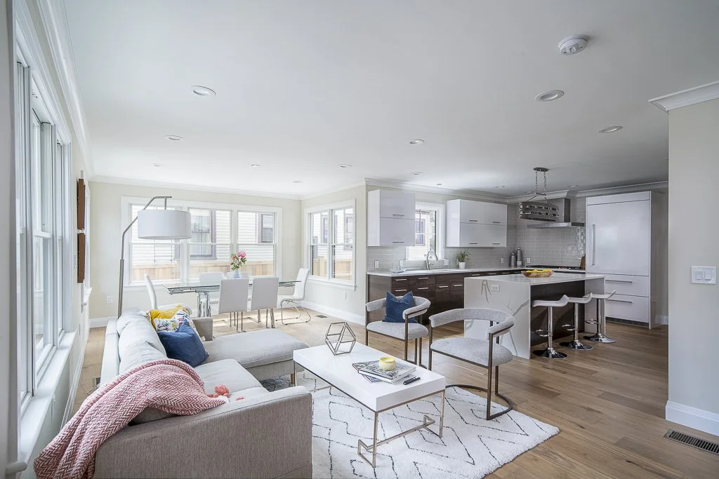 Open-concept living room and kitchen with white and gray decor, hardwood floors, large windows, and modern furniture.