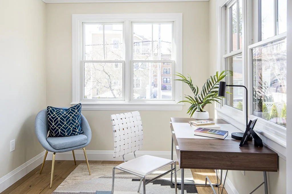 A bright home office with a window view, a desk with a computer, books, and a plant, along with a chair and a cozy armchair with a pillow.