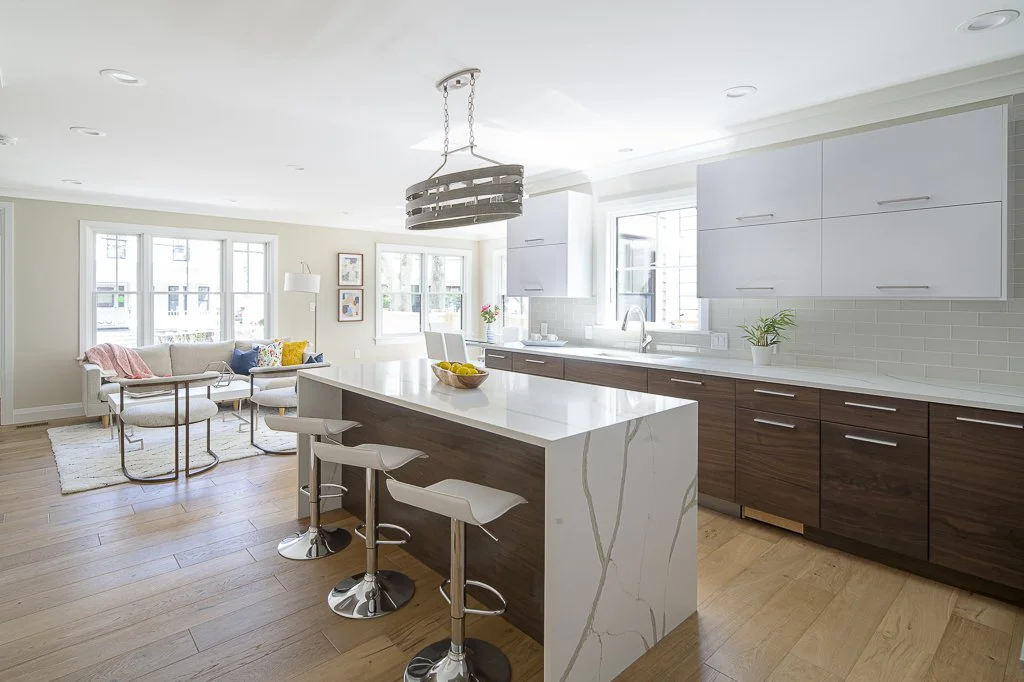 Bright open-concept kitchen and living room with white upper cabinets, wood lower cabinets, and a white island with bar stools. The living area has a white sofa, colorful pillows, a side table, and large windows letting in natural light.