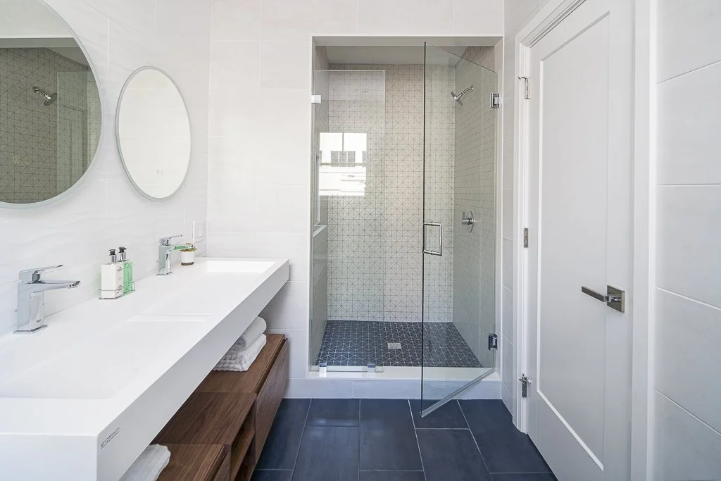 Modern bathroom with a white double sink vanity, wooden storage cabinet, frameless glass shower with tiled walls and floor, and two round mirrors above the sinks.