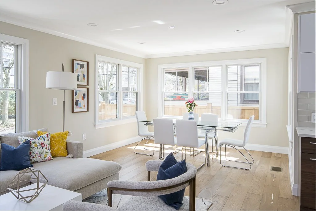 Bright living and dining area with large windows, white chairs around a glass table with a vase of pink flowers, light-colored sofa with colorful pillows, and modern decor.