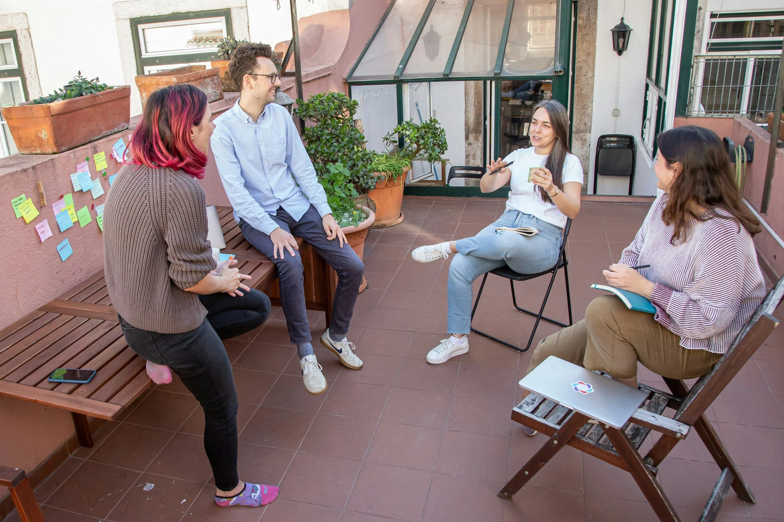 Group of five young adults on a rooftop terrace engaging in conversation, some sitting and some standing, with plants and colorful sticky notes on the wall.