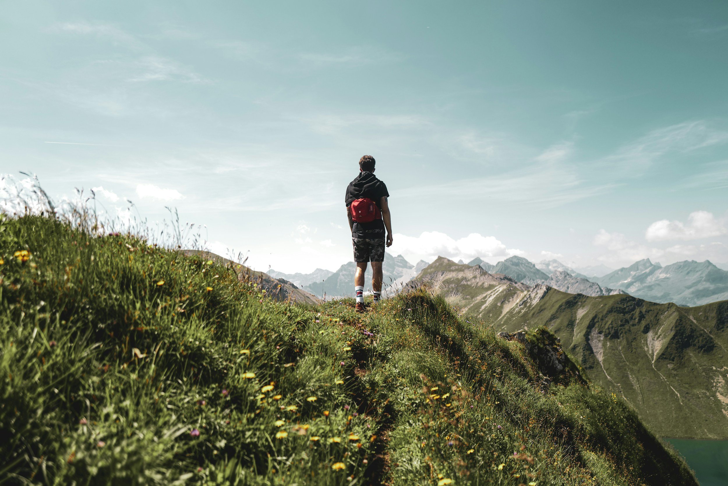 A person with a backpack walking on a grassy mountain trail surrounded by wildflowers, with mountains in the distance and a blue sky with wispy clouds.