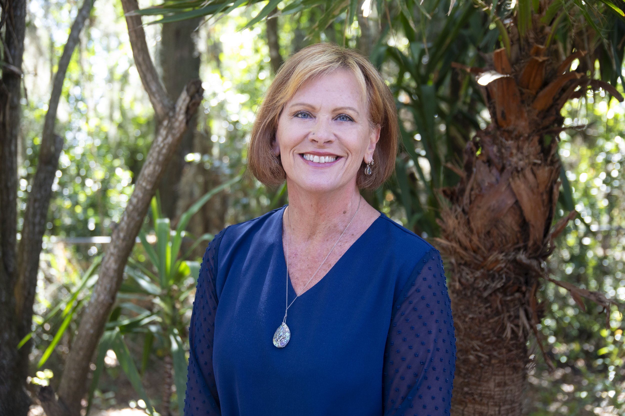 A woman with shoulder-length red hair, wearing a blue dress and jewelry, standing outdoors in front of trees and greenery, smiling at the camera.