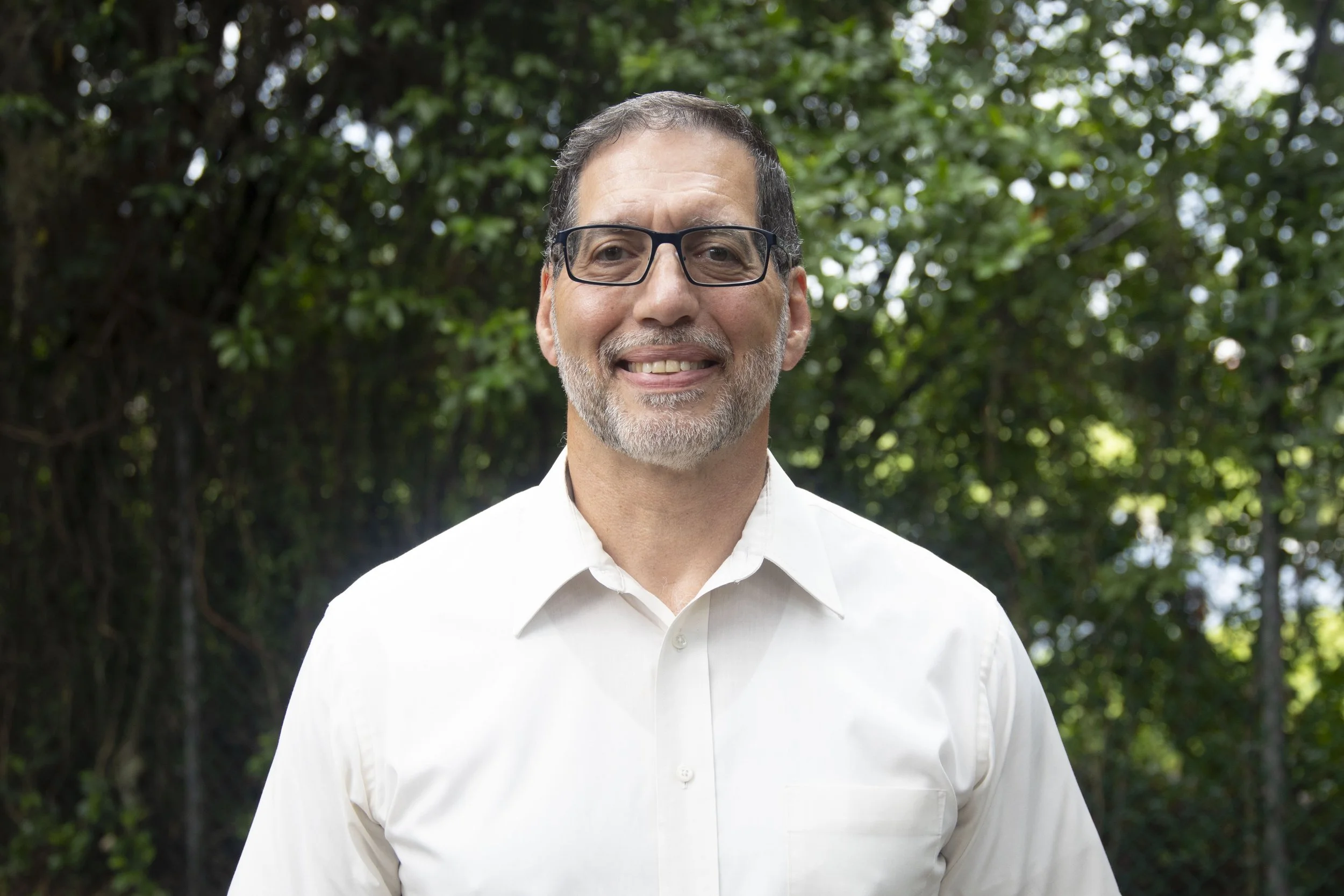 A middle-aged man with dark hair, glasses, and a beard, smiling outdoors in front of green trees, wearing a white shirt.
