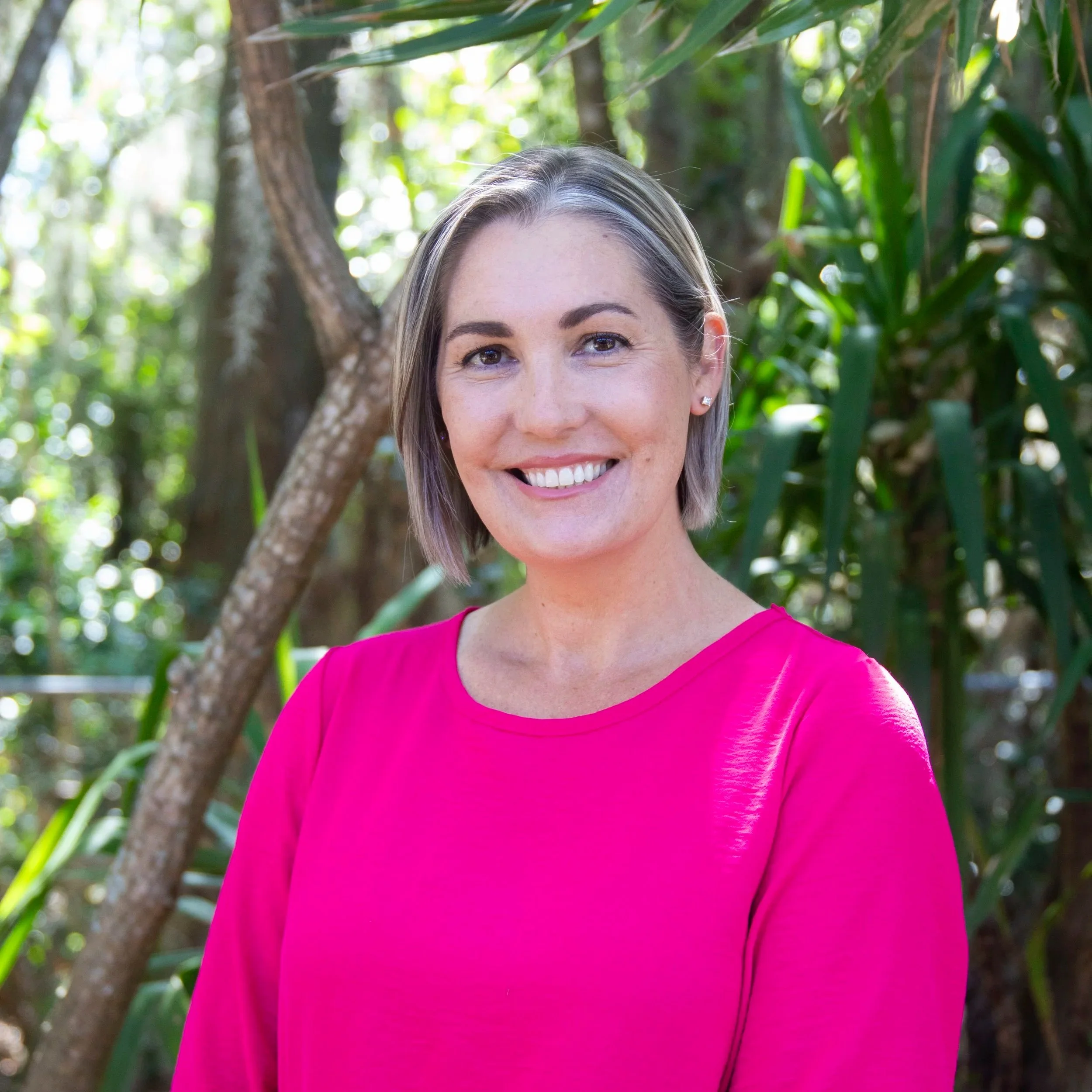 A woman with short, light-colored hair smiling in a pink top, standing outdoors with green trees and foliage in the background.