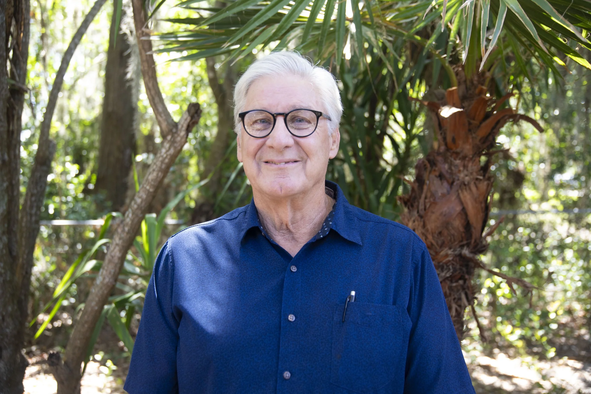 A smiling elderly man with gray hair and glasses wearing a blue shirt, standing outdoors in a lush green area with trees and sunlight filtering through leaves.