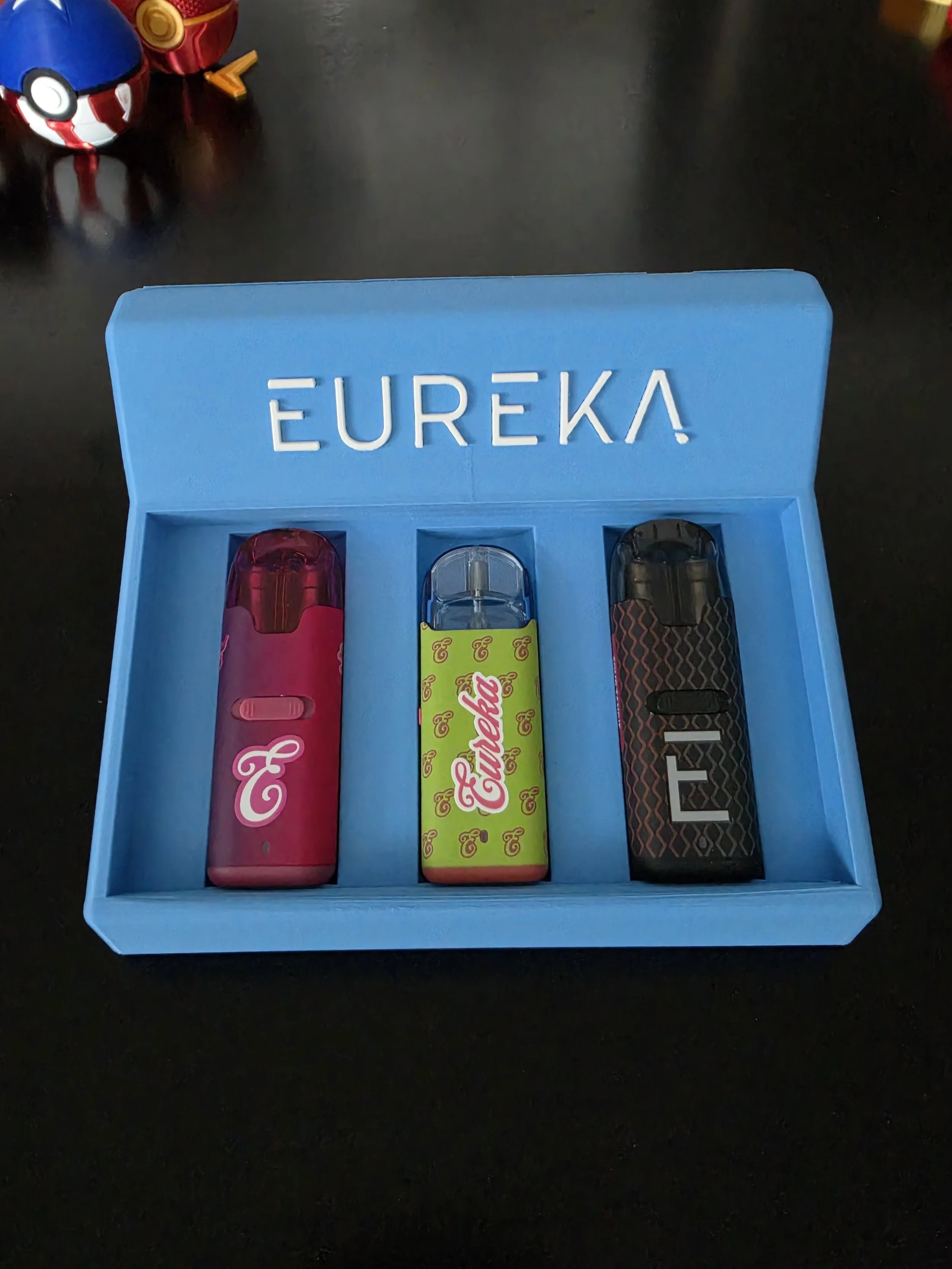Blue box labeled 'EUREKA' contains three Nintendo Switch game cartridges on a black surface, with Pokémon themed Poké Balls partially visible in the background.